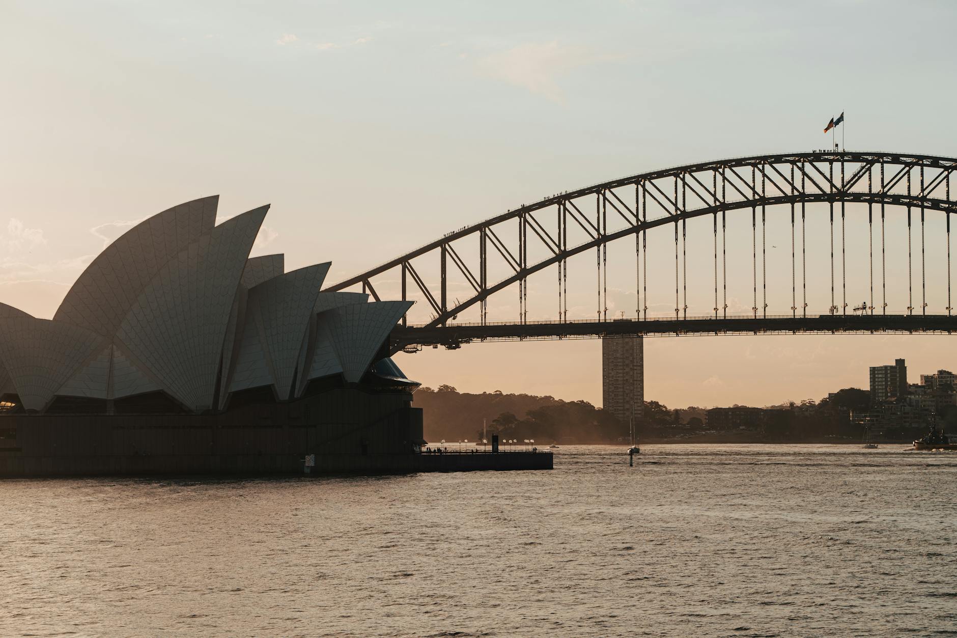 The sails of the Sydney Opera House and the Harbour Bridge silhouetted against a soft golden sunset sky over calm harbour water