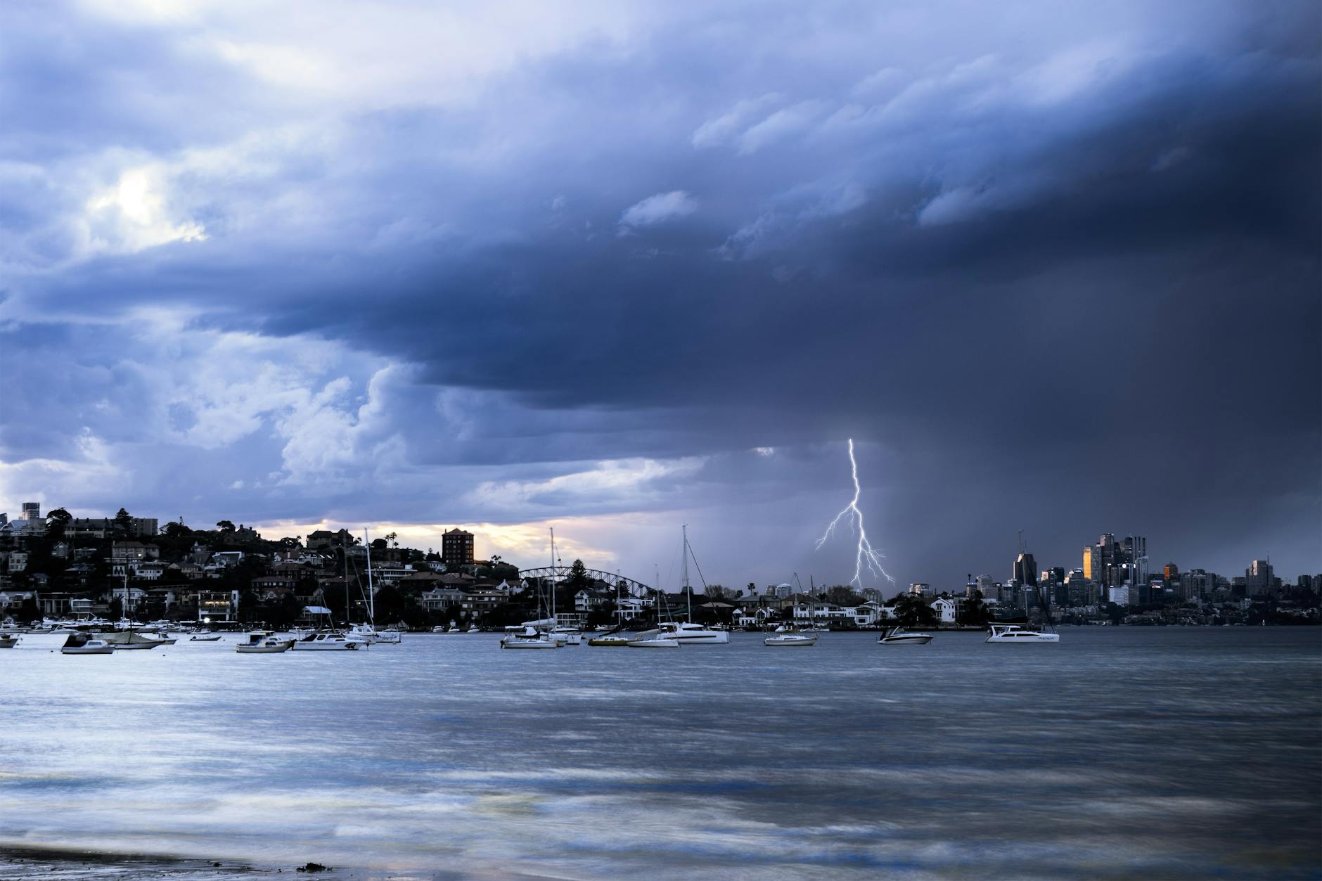 Dark storm clouds over the eastern harbour with a lightning bolt striking behind moored boats and the city skyline on the right