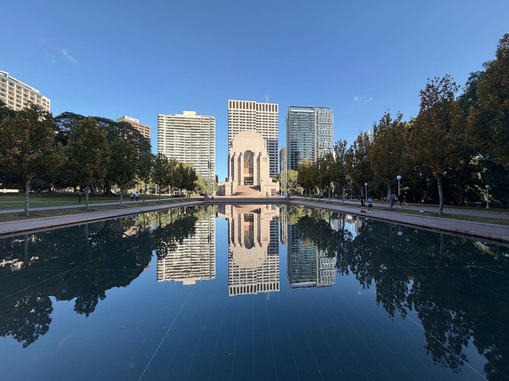 The Art Deco Anzac Memorial at the end of the long Pool of Reflection in Hyde Park, with autumn trees along both sides and city towers behind under a clear blue sky