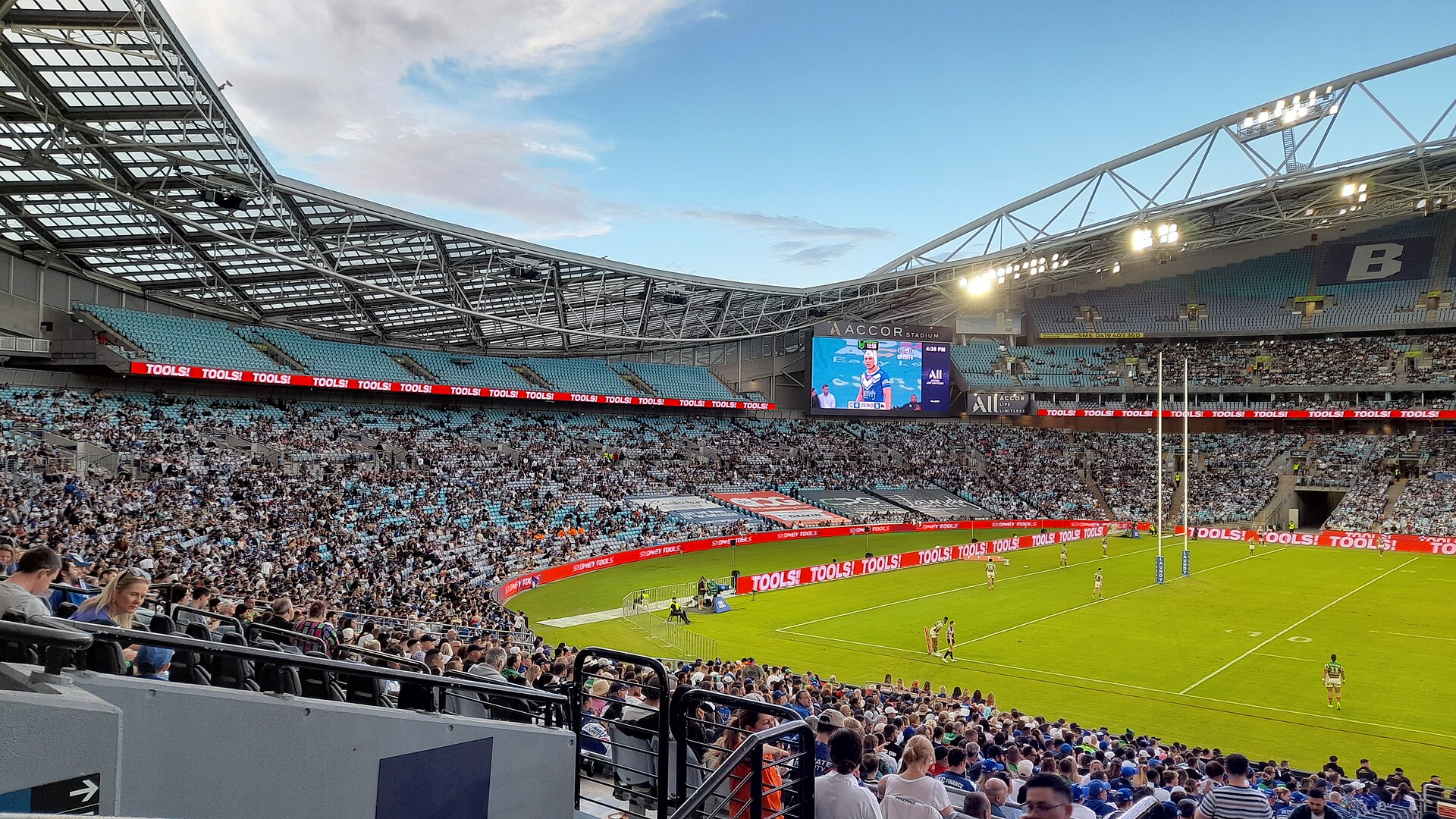 Inside Accor Stadium at Sydney Olympic Park during a match, with a large crowd in the lower tier, the curved roof overhead and players on the green field