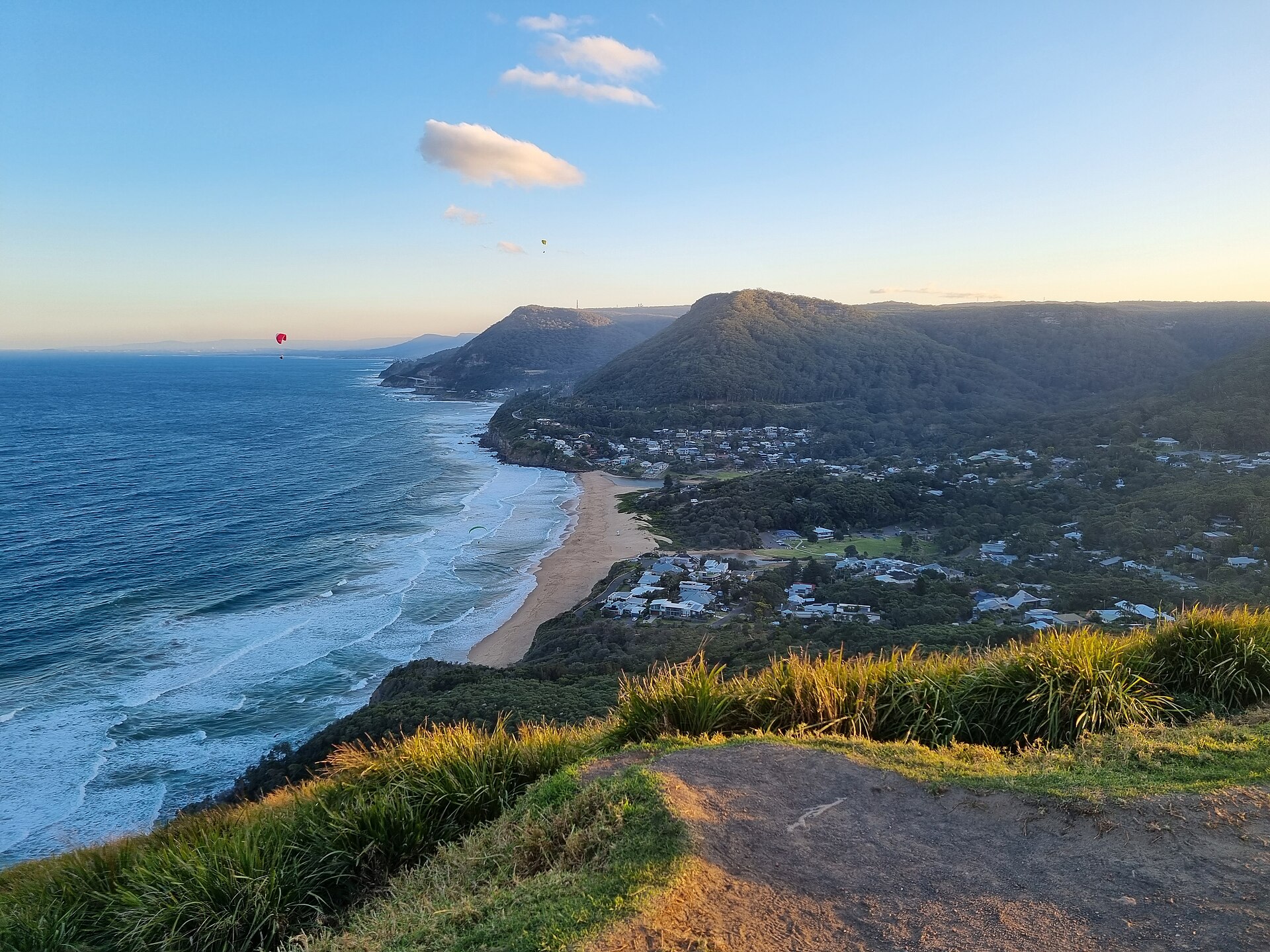 View south from Bald Hill Lookout over Stanwell Park beach and village, with forested escarpment, the coast road curving away and paragliders in the evening sky