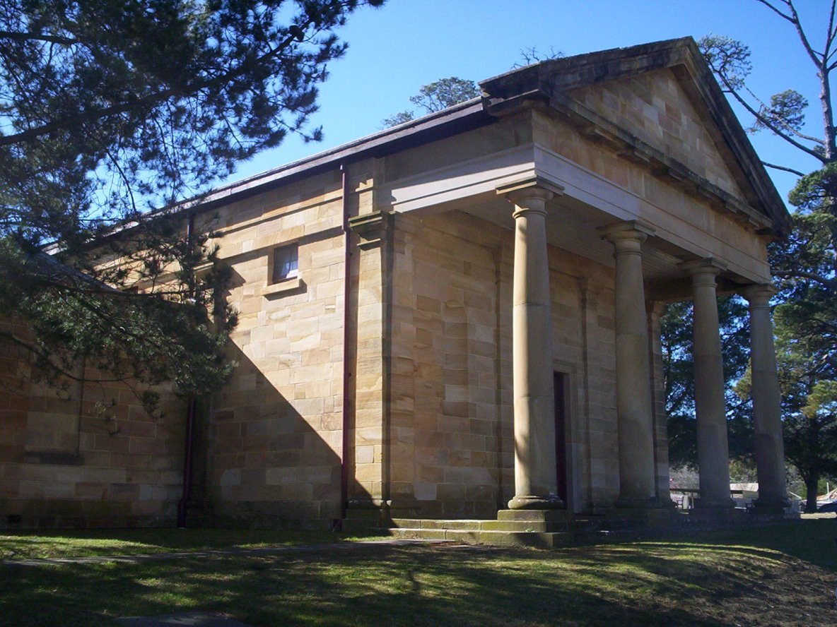 The sandstone Berrima Courthouse with its columned portico and pediment, partly shaded by pine trees under a clear blue sky
