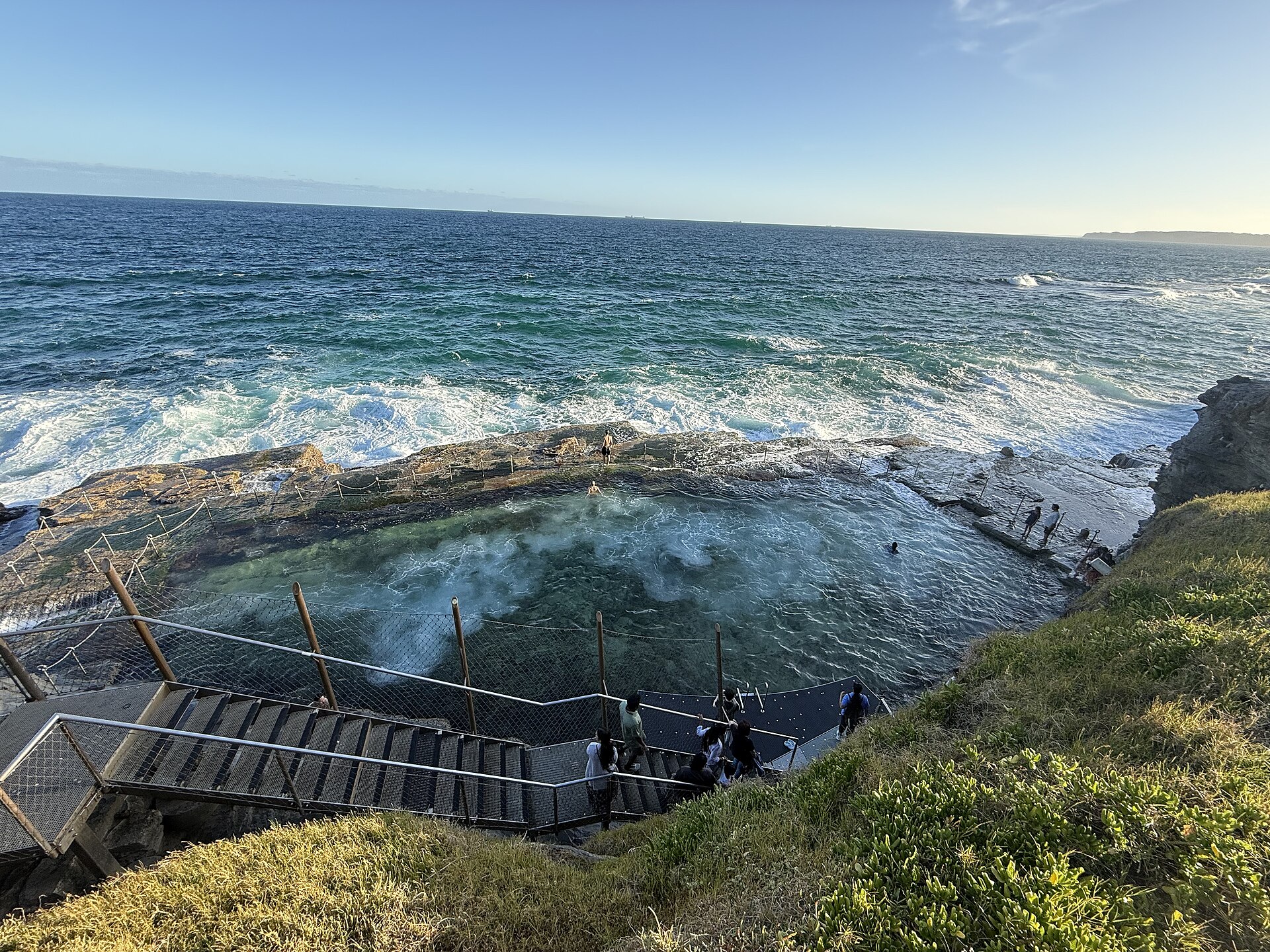 Swimmers in the Bogey Hole, a rectangular pool cut into a rock platform, with surf breaking over its outer edge and a steel staircase leading down from the grassy cliff