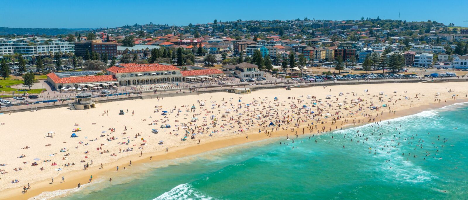 Aerial view of Bondi Beach on a hot, clear summer day, with crowds on the sand, swimmers in the surf and the Bondi Pavilion behind