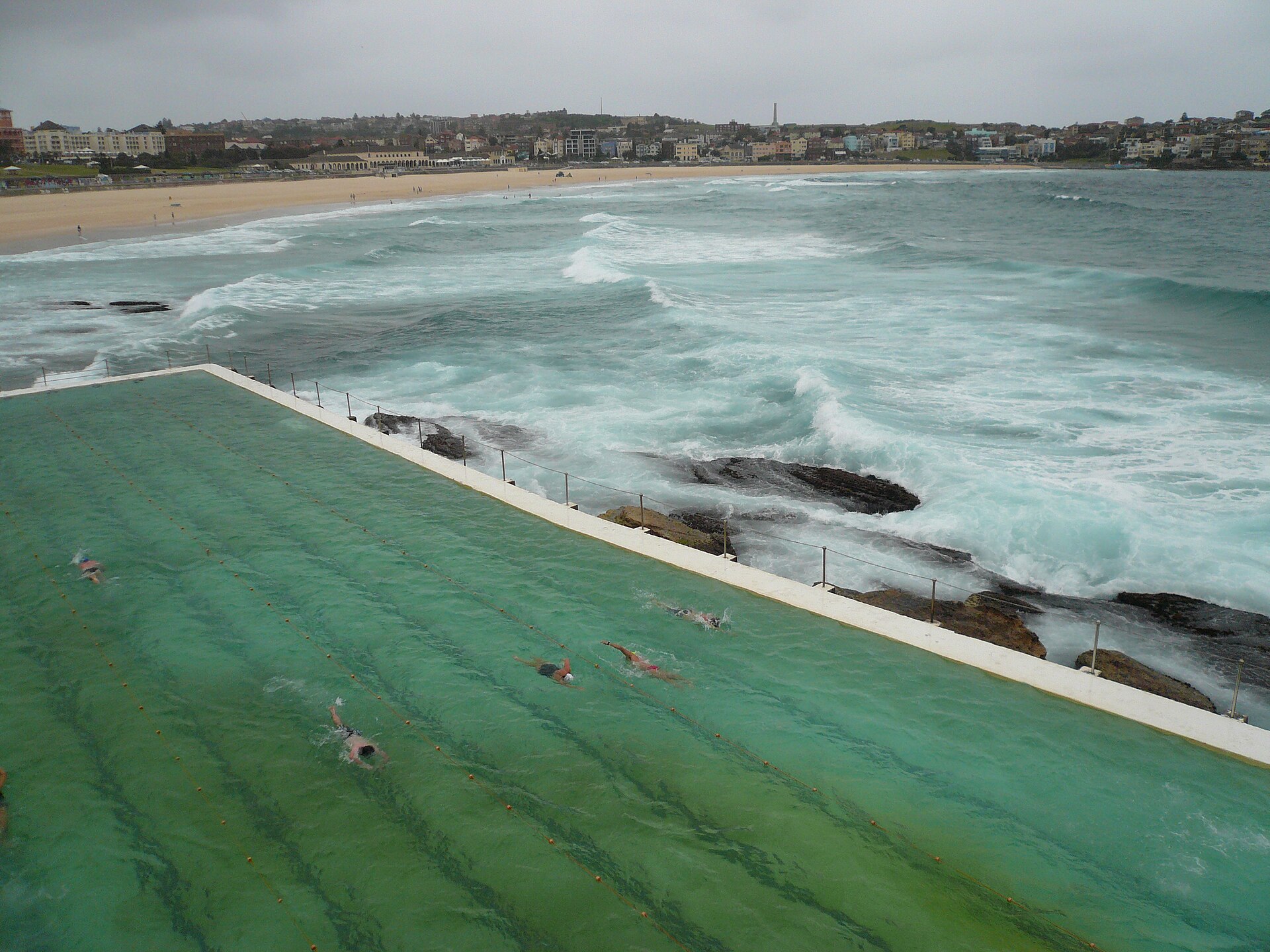 Swimmers doing laps in the Bondi Icebergs ocean pool on a grey day, with rough surf breaking over the rocks beside it and Bondi Beach curving away behind