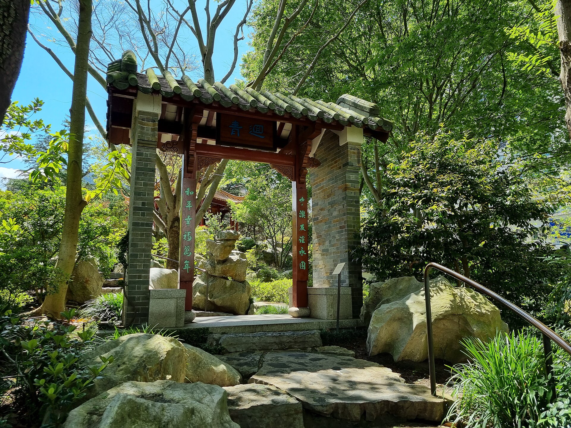 A gateway with a green-glazed tiled roof, red timber posts and panels of Chinese calligraphy, set among trees, rocks and ferns on a paved path