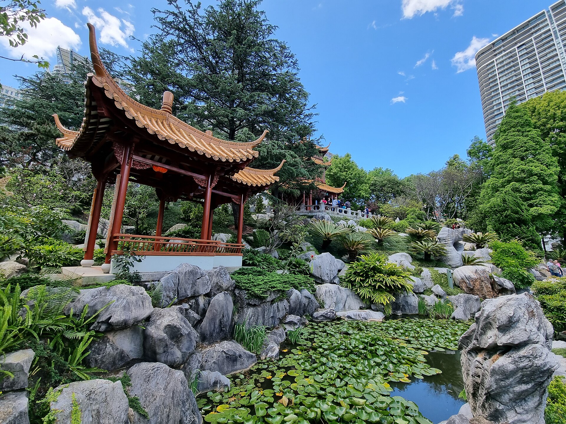 A red timber pavilion with sharply upswept tiled eaves standing on a rockery of grey stones above a lily pond, with trees and an apartment tower in the background