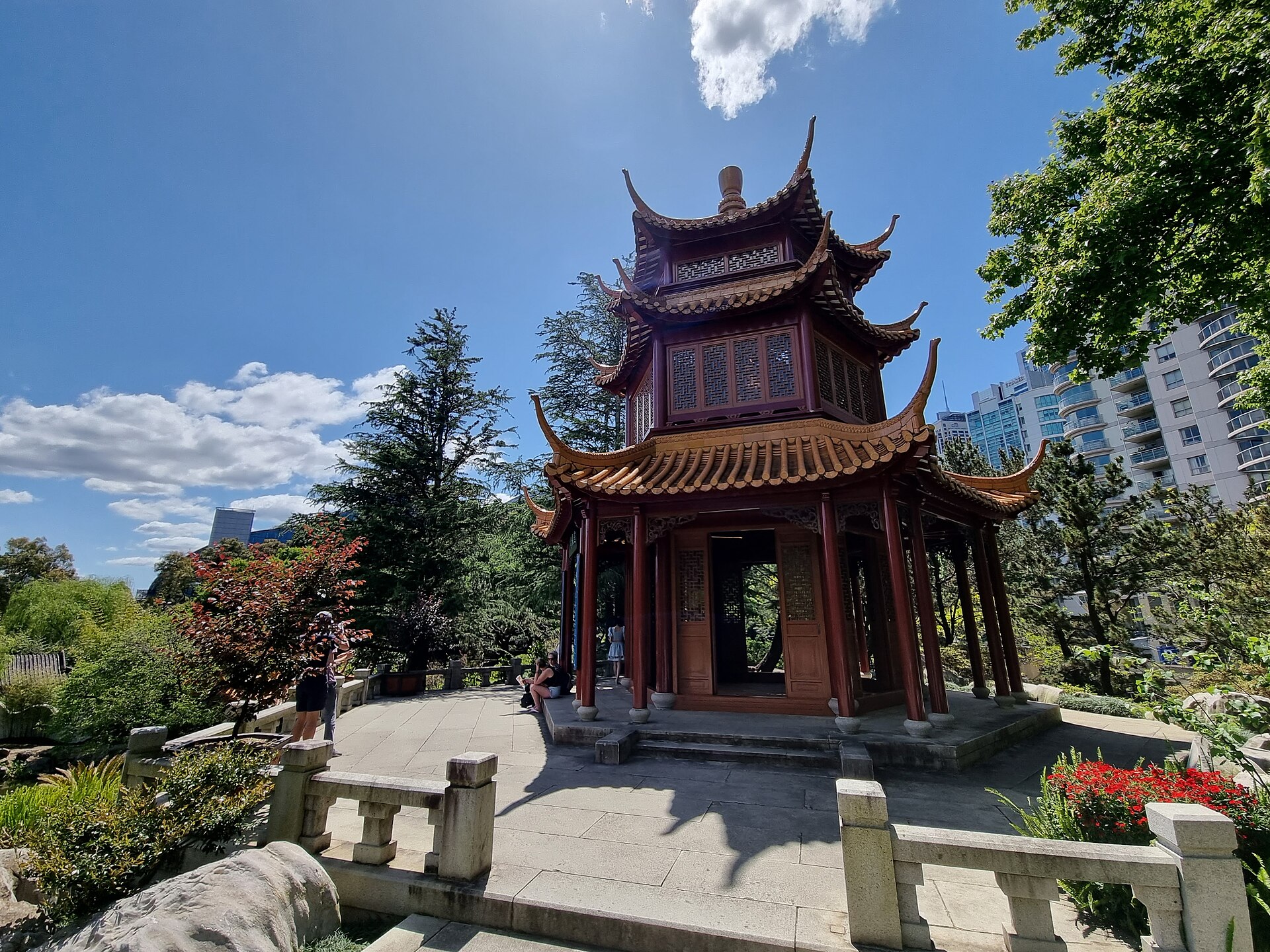 A tall tiered Chinese pavilion with three layers of upswept brown-tiled roofs and red timber columns, standing on a paved stone terrace with a carved balustrade, trees and apartment towers behind