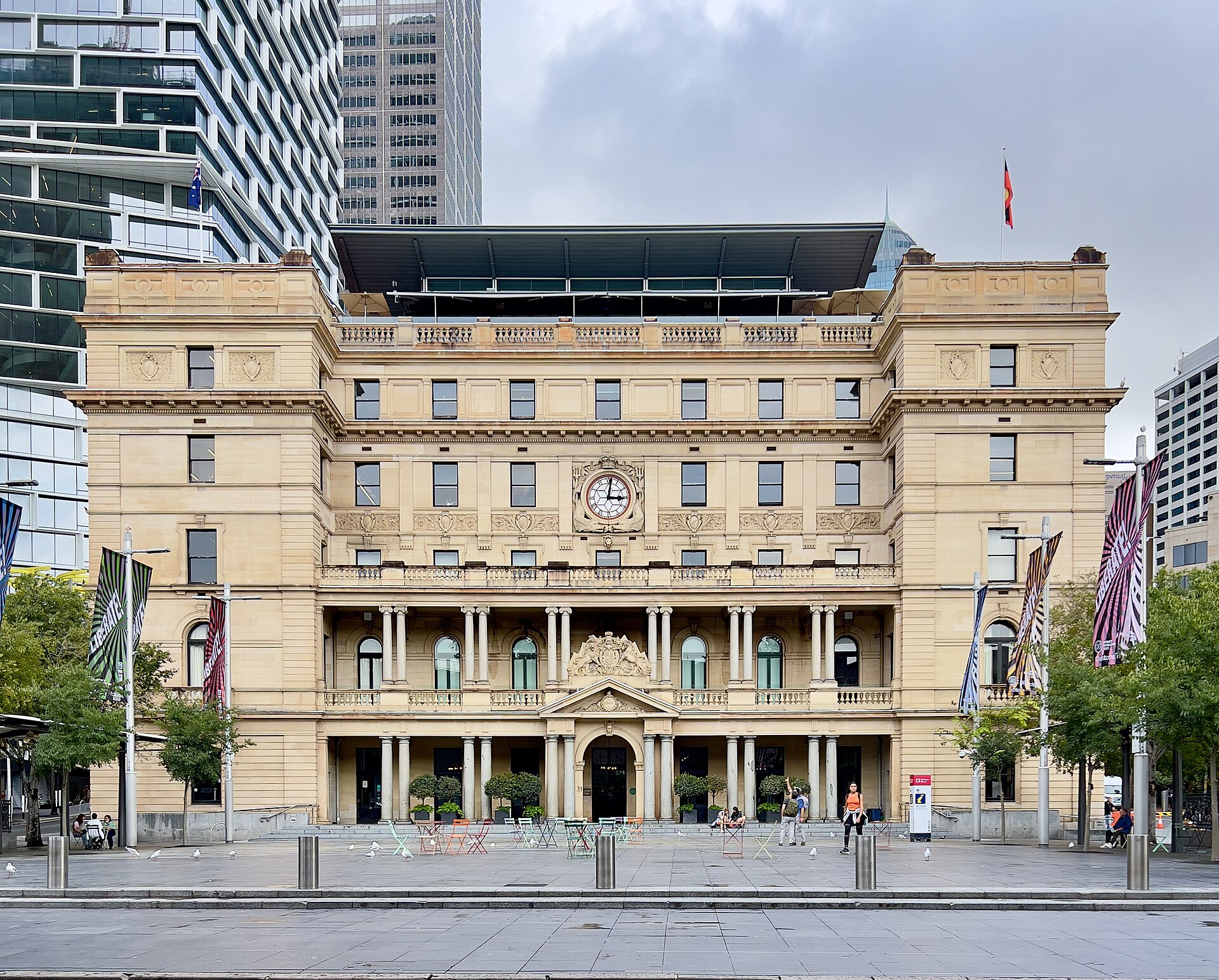 The sandstone facade of Customs House at Circular Quay, with a central clock, columns and carved coat of arms, and a paved square in front