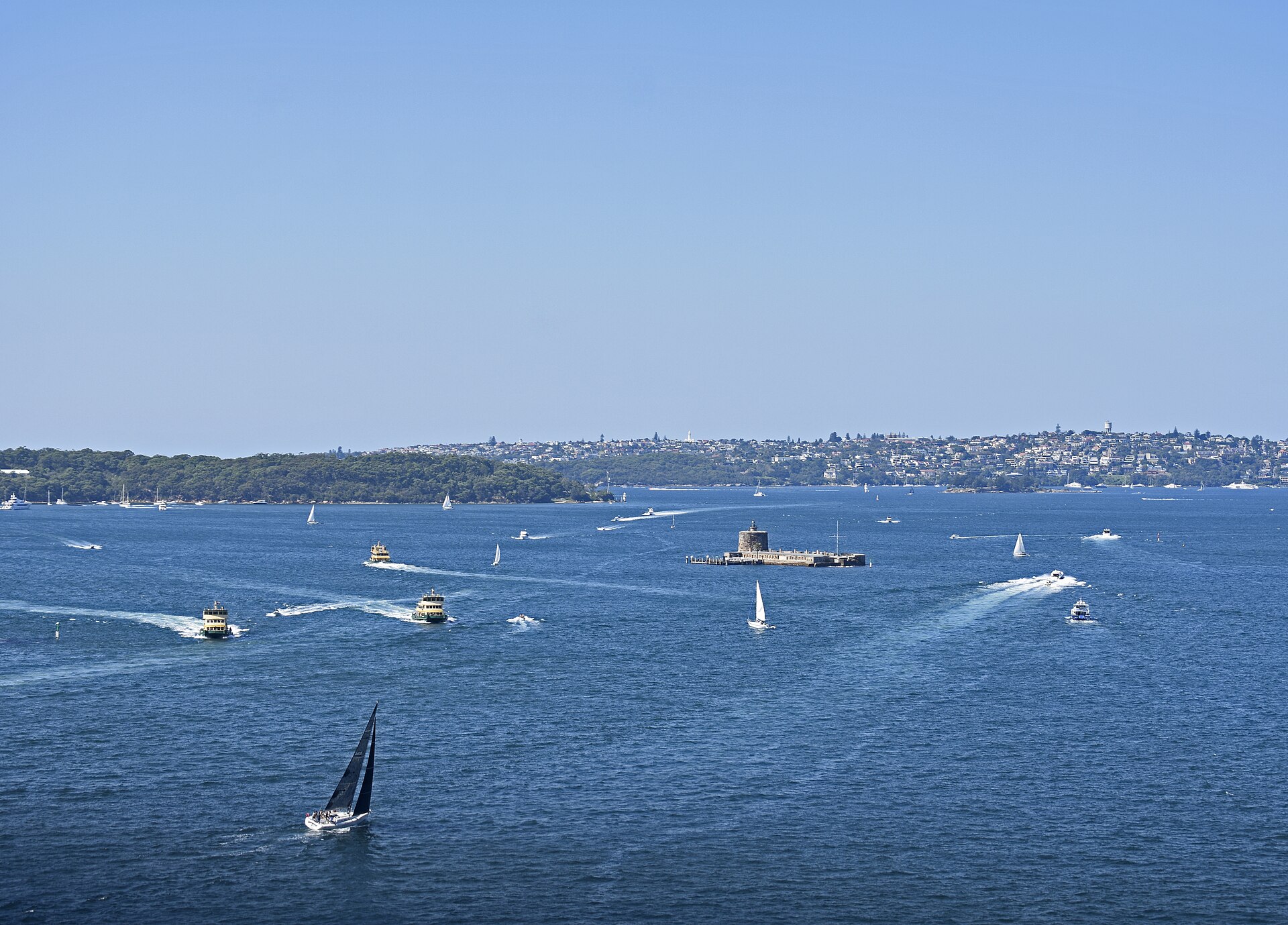 Fort Denison, a small stone fort with a round tower on a low island in the middle of Sydney Harbour, surrounded by ferries, yachts and motorboats on bright blue water