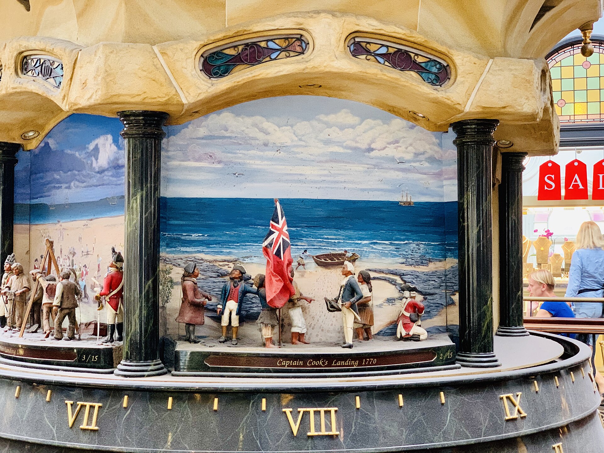 A detailed diorama on the Great Australian Clock showing Captain Cook's landing in 1770, with painted figures, a red ensign and a beach scene above a band of Roman numerals