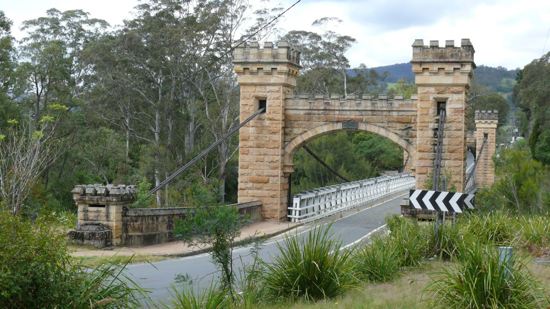 Hampden Bridge at Kangaroo Valley, a suspension bridge with two castellated sandstone towers joined by an arch, white railings and a narrow road deck among gum trees