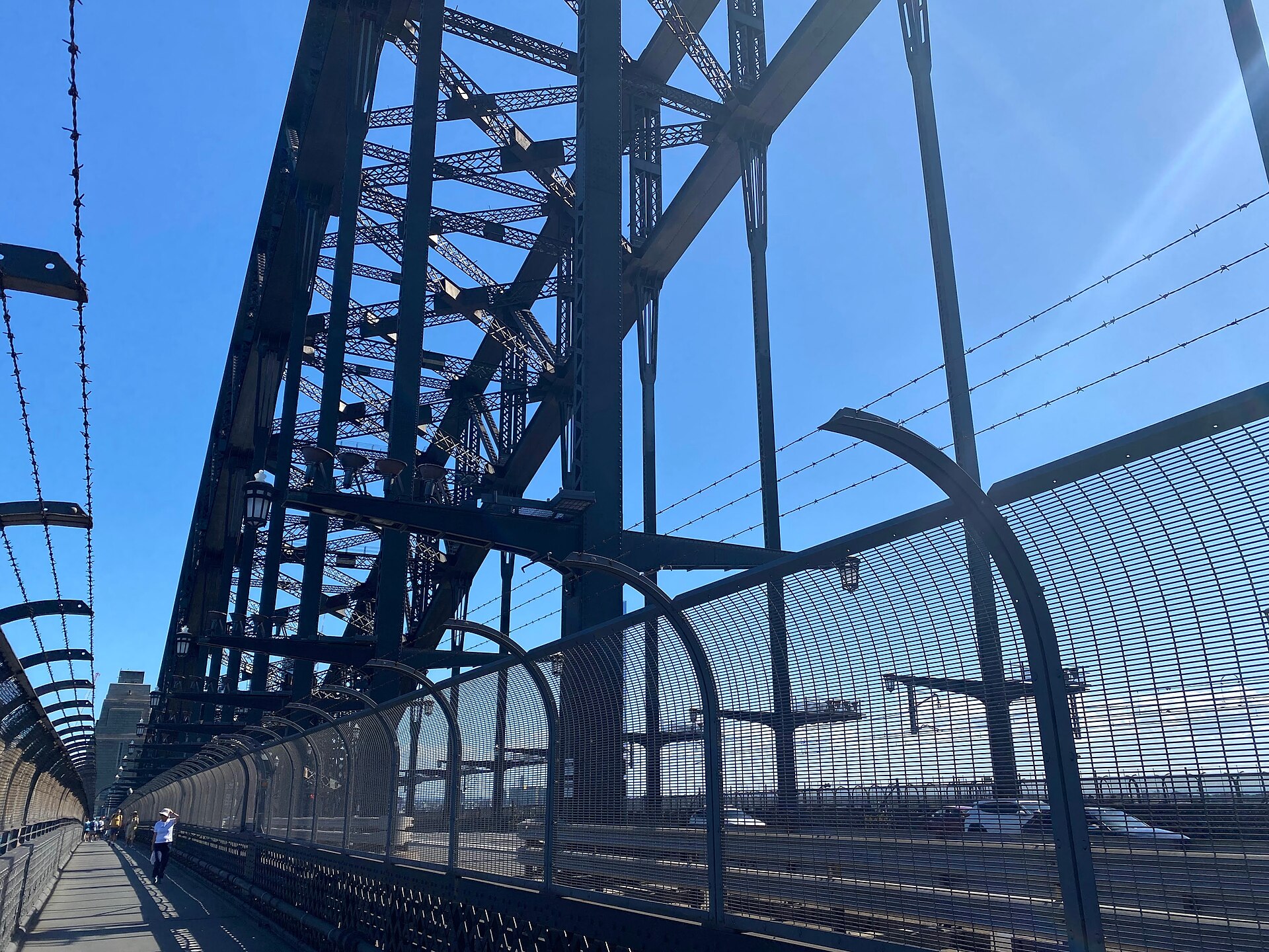The steel arch of the Sydney Harbour Bridge rising over the pedestrian footpath, with curved safety fencing, a walker in the distance and traffic on the roadway beside