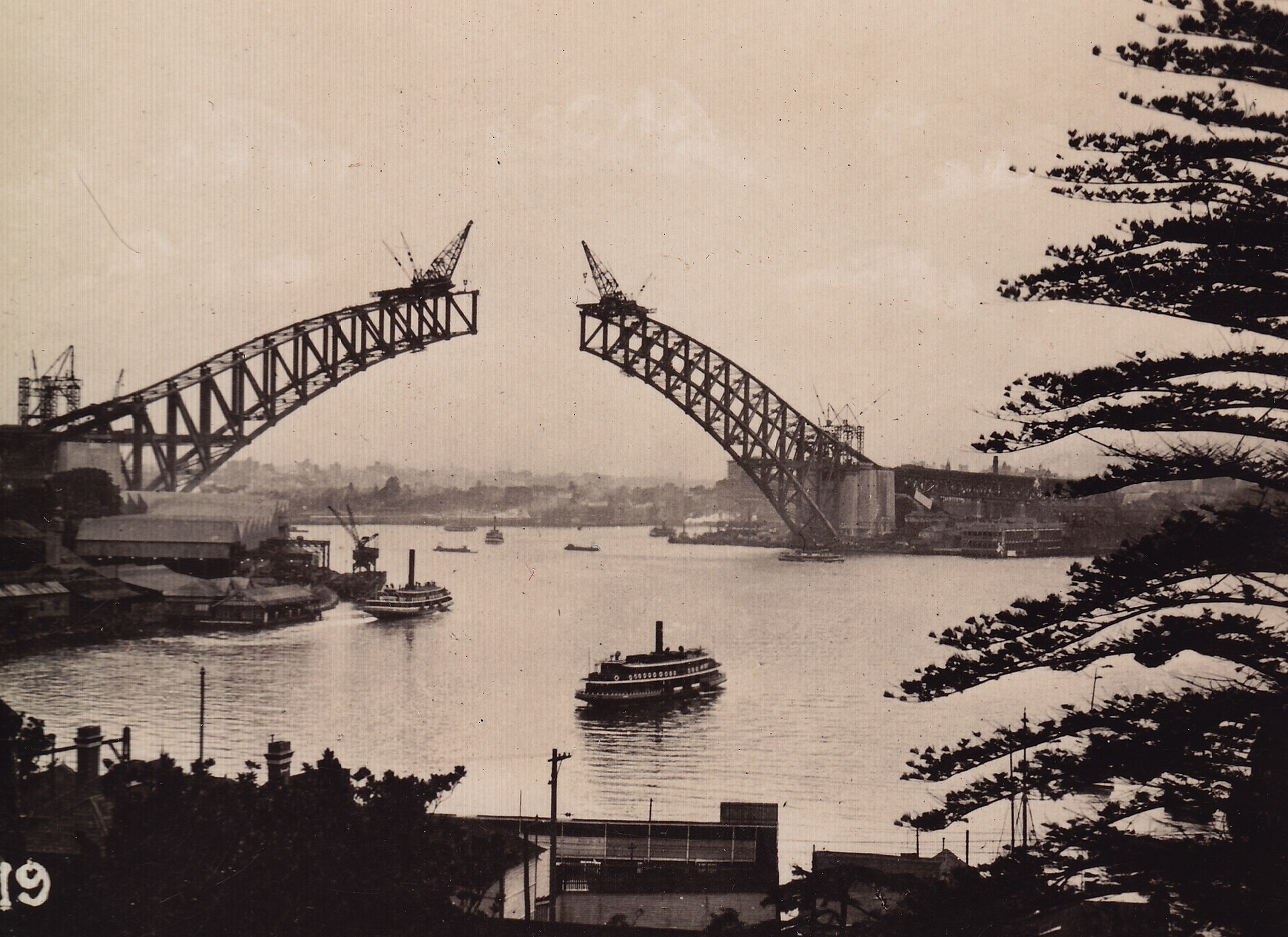 A sepia photograph of the two unfinished halves of the Harbour Bridge arch reaching towards each other across the harbour, with cranes on top, a ferry below and pine trees in the foreground, around 1930