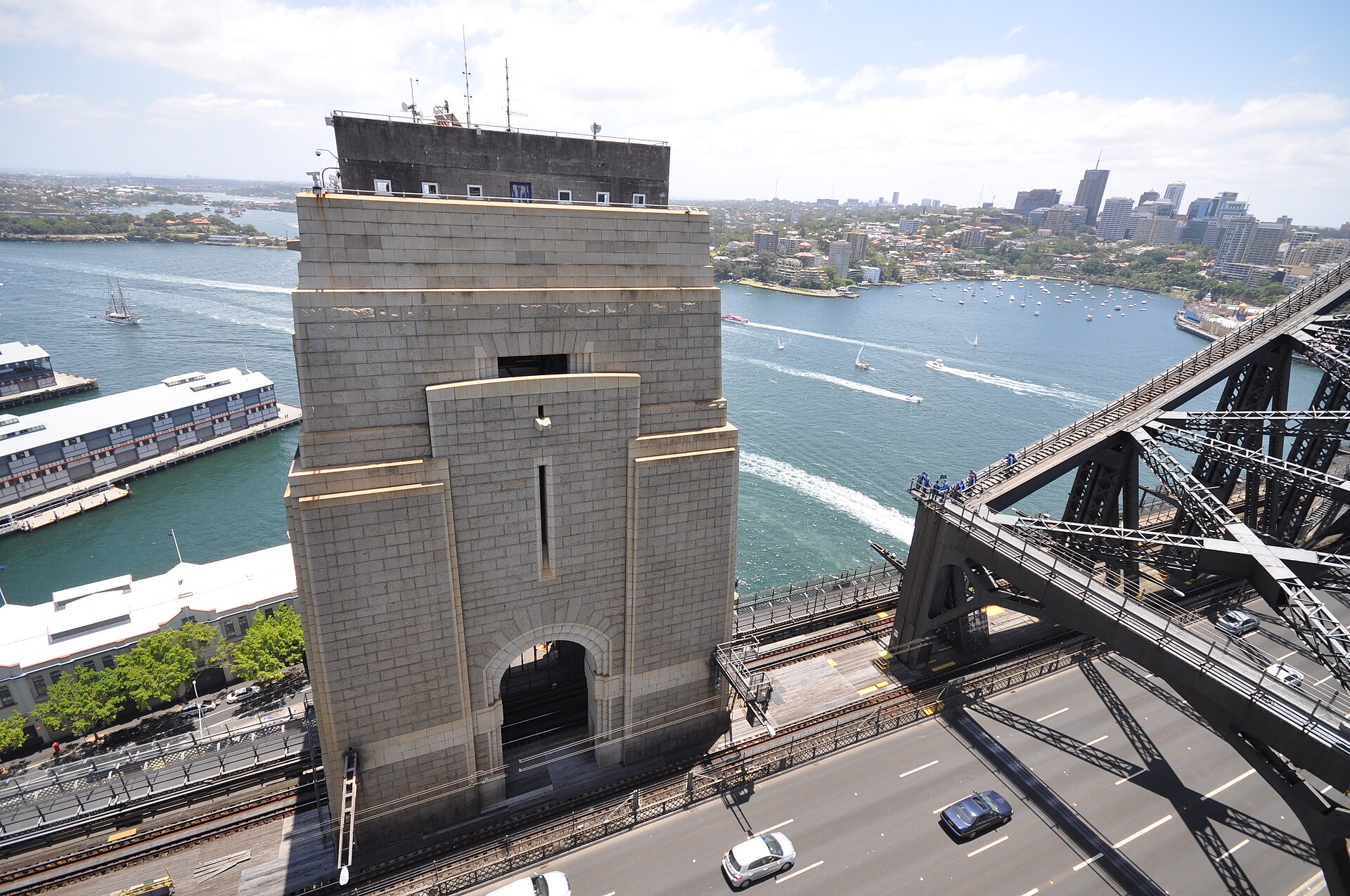 Looking down from the south-east pylon to the granite-faced south-western pylon of the Harbour Bridge, with the roadway and railway below, climbers on the arch and the harbour behind