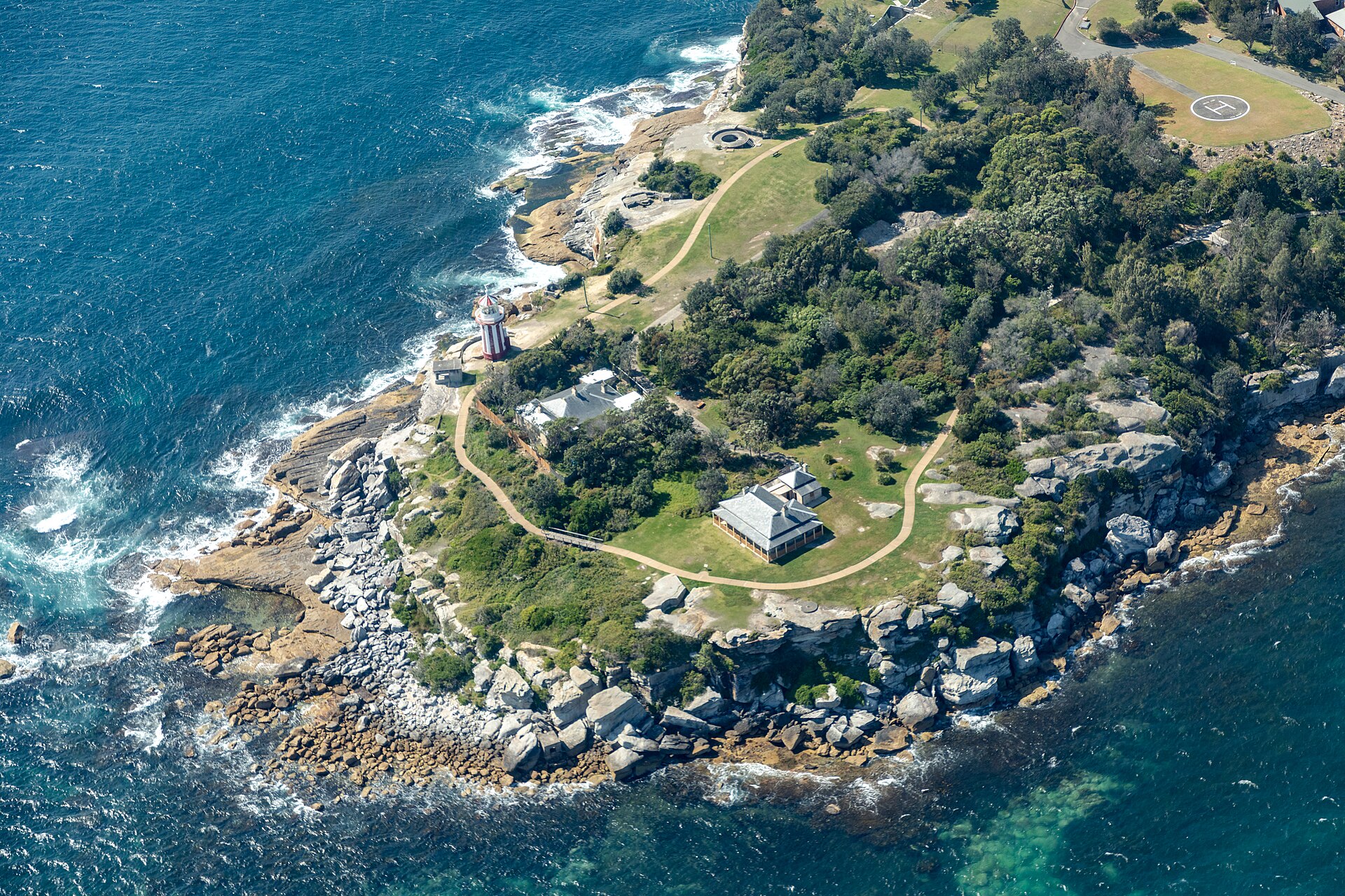 The red-and-white striped Hornby Lighthouse on the rocky tip of South Head, seen from above, with walking paths, old cottages and the blue ocean around it