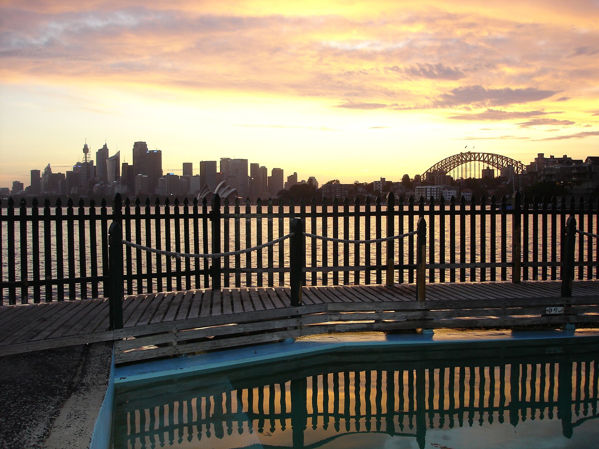 Maccallum Pool at Cremorne Point at sunset, with its timber deck and picket fence in silhouette and the Sydney skyline and Harbour Bridge across the glowing water