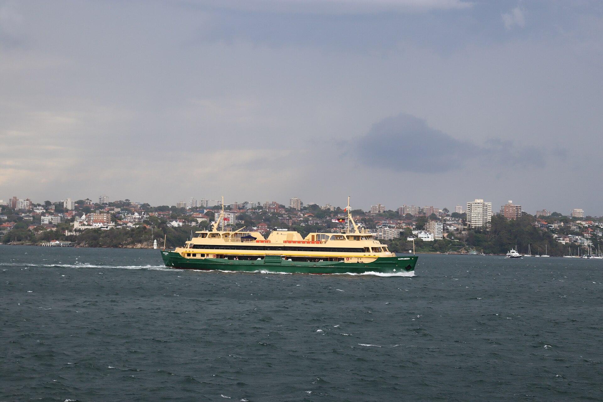 A green-and-cream Freshwater-class Manly ferry crossing the harbour under a grey sky, with the eastern suburbs' apartments and houses on the shore behind