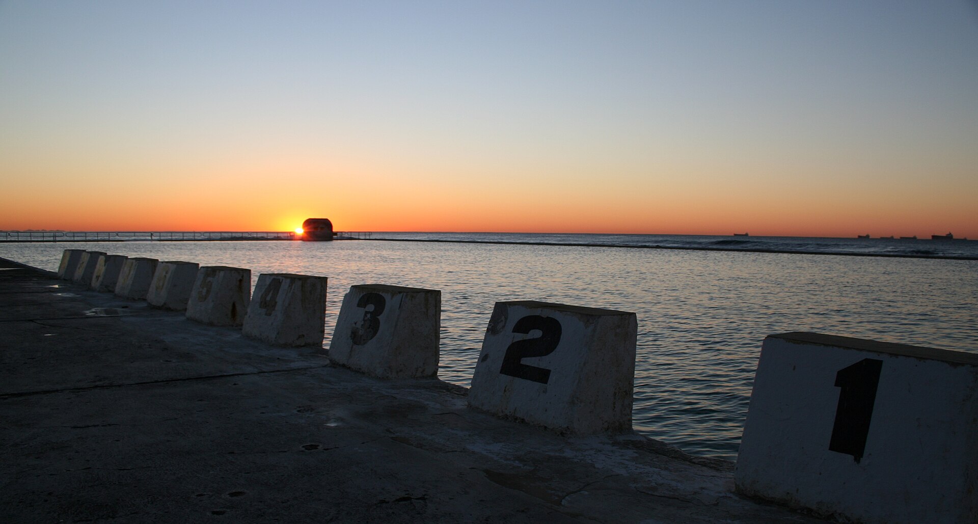 Numbered concrete starting blocks along the edge of Merewether Ocean Baths at sunrise, with the sun rising behind the far wall and ships on the horizon