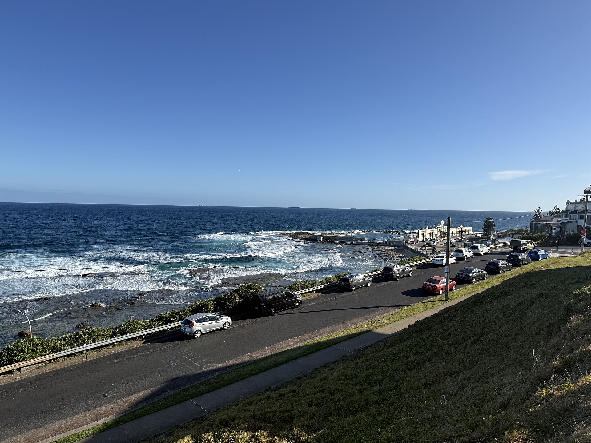 Newcastle Ocean Baths and its cream pavilion beside a rock shelf and breaking surf, with parked cars along the road above and ships on the horizon