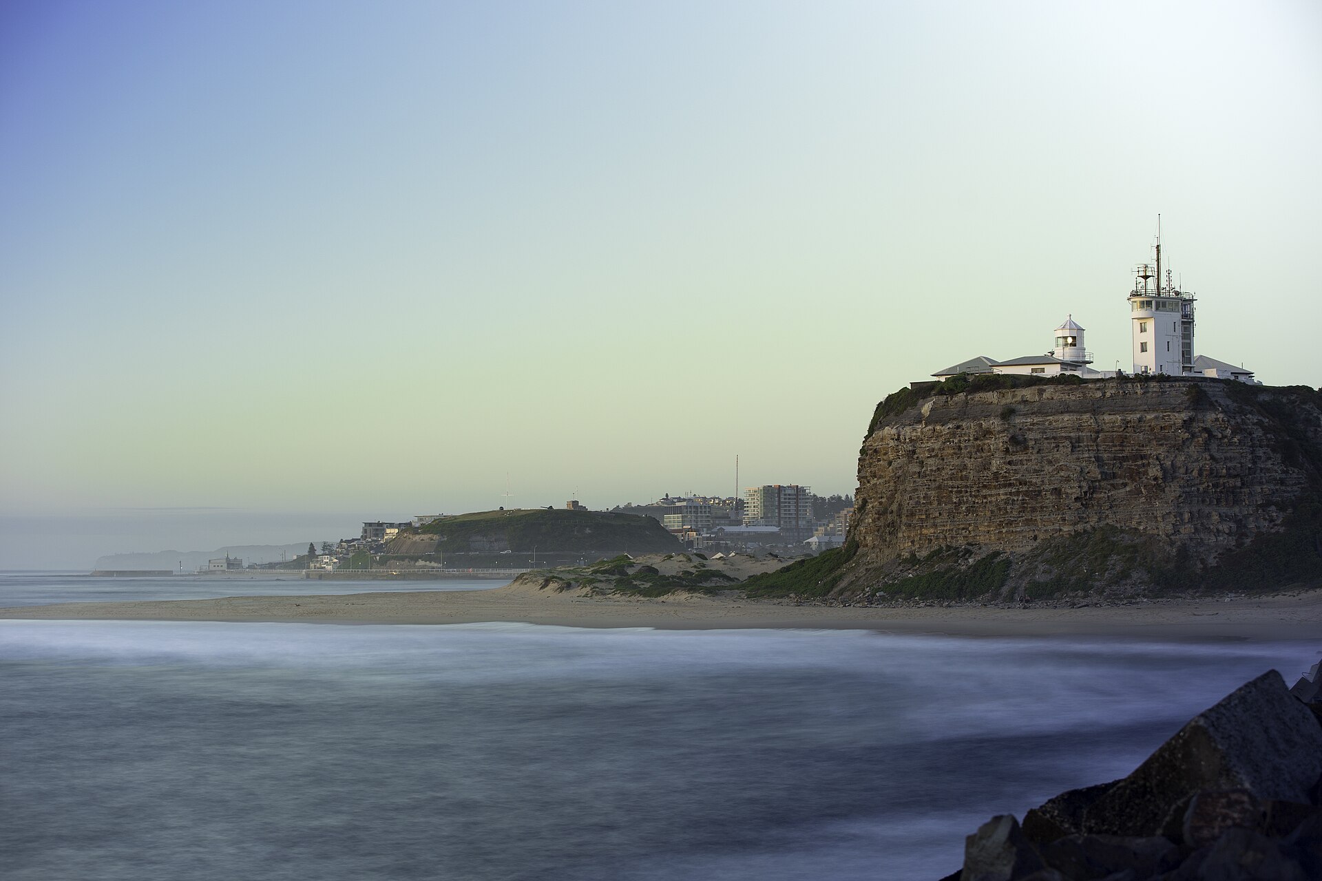 Nobbys Head Lighthouse and signal station on top of a sheer layered cliff at dusk, with the beach and the Fort Scratchley hill behind