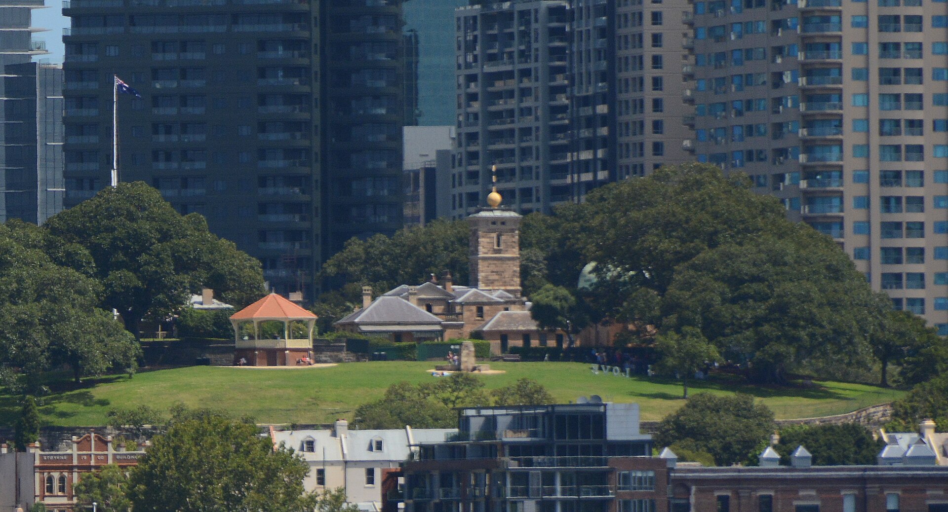 Observatory Hill seen from a distance, a grassy rise with a white bandstand and the sandstone Sydney Observatory with its tower among large fig trees, and apartment and office towers behind