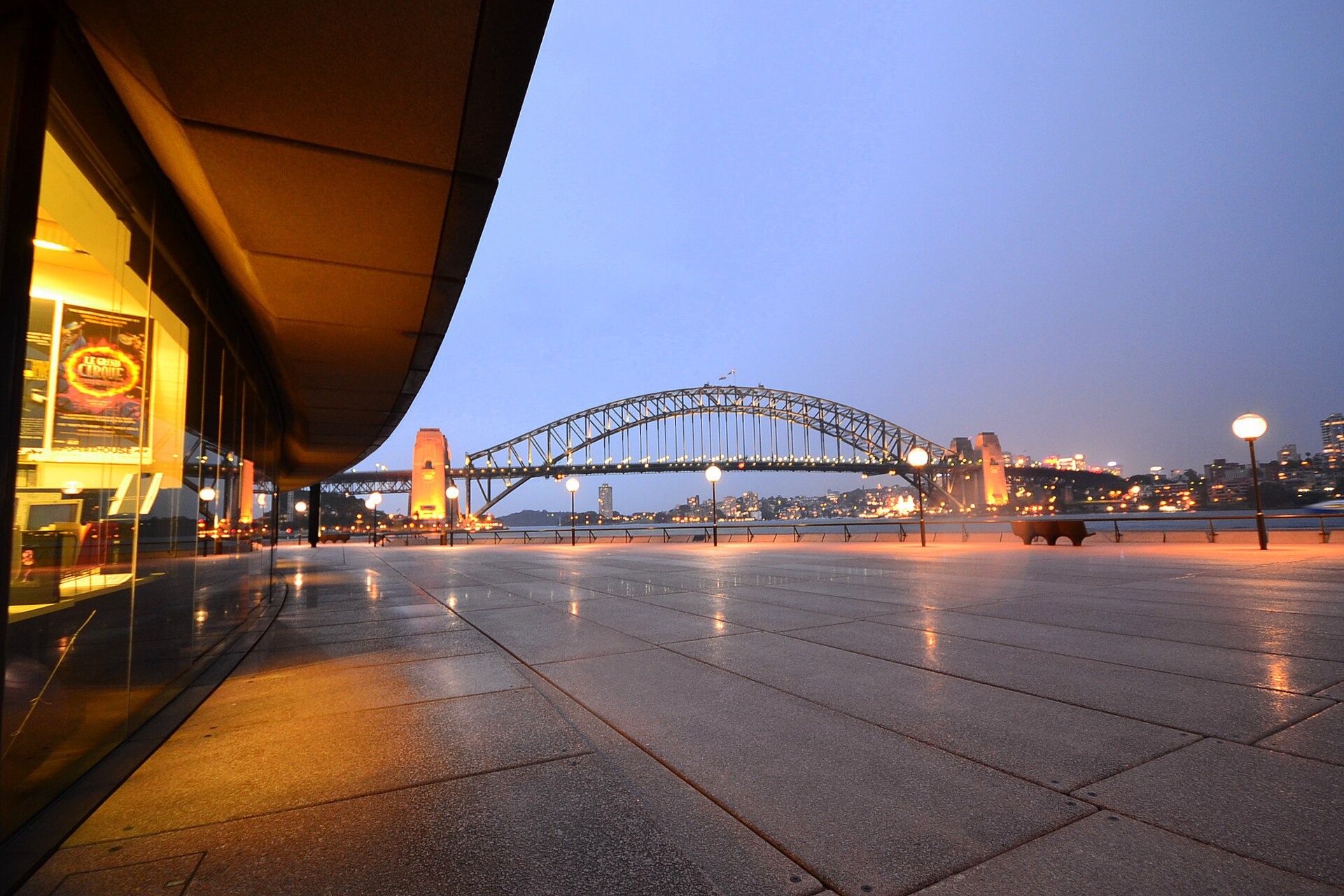 The Opera House's curved lower concourse at dusk, its lights reflected on the wet paving, with the Harbour Bridge lit up across the water