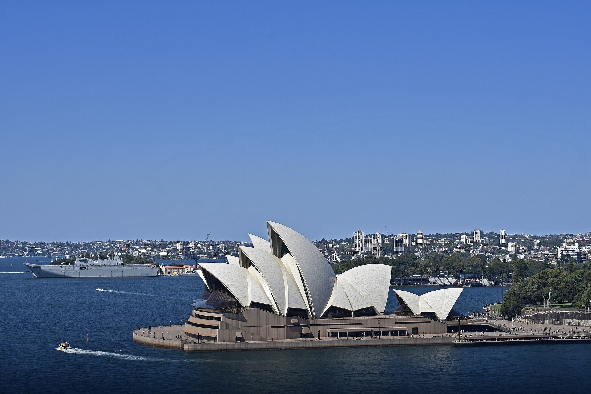 The Sydney Opera House and the harbour seen from high on the Harbour Bridge pylon, with a grey warship moored at Garden Island behind and small boats on the blue water