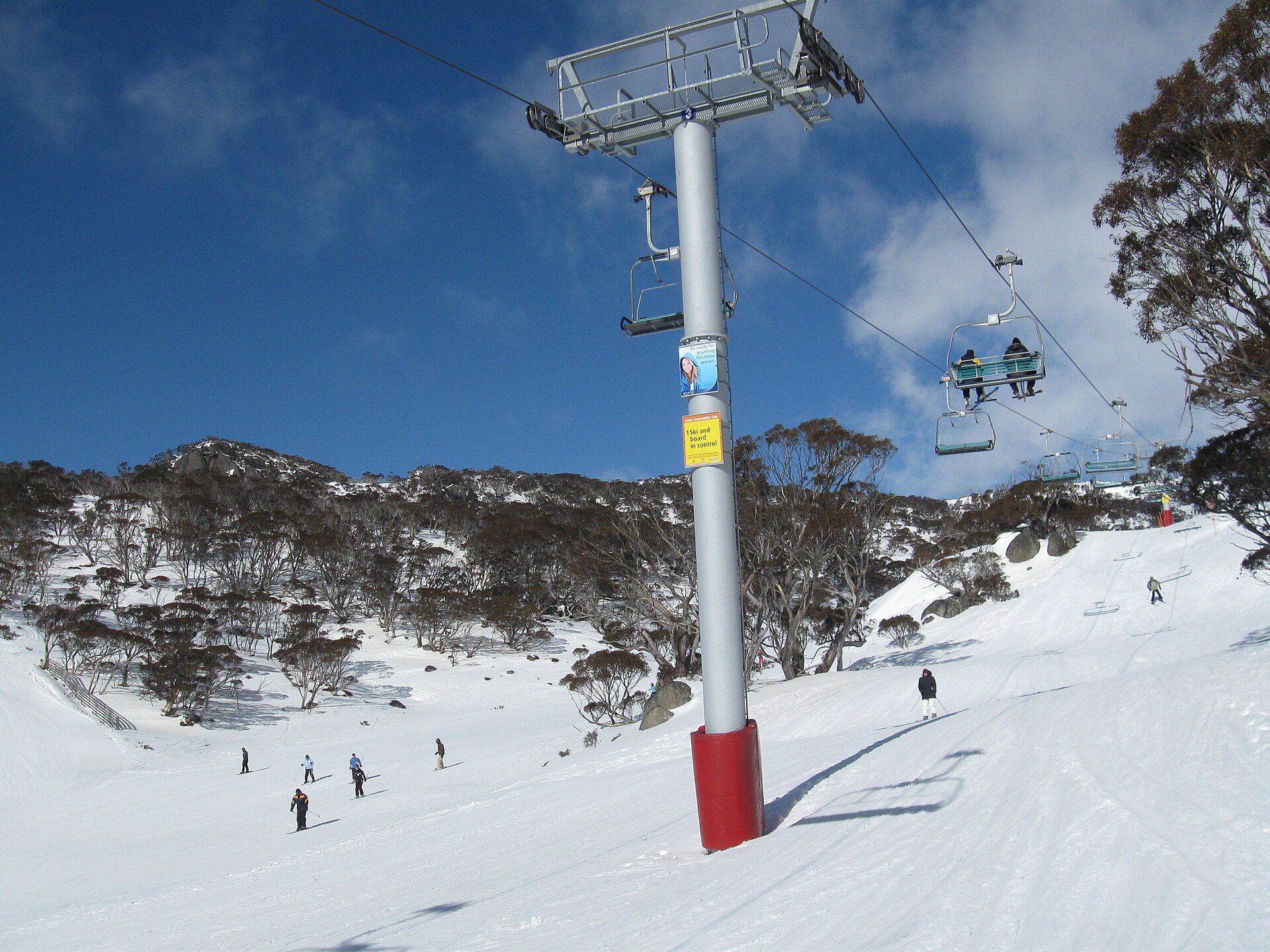 A chairlift rising over a snowy run at Perisher, with skiers on the slope, snow gums on the hillside and a deep blue sky