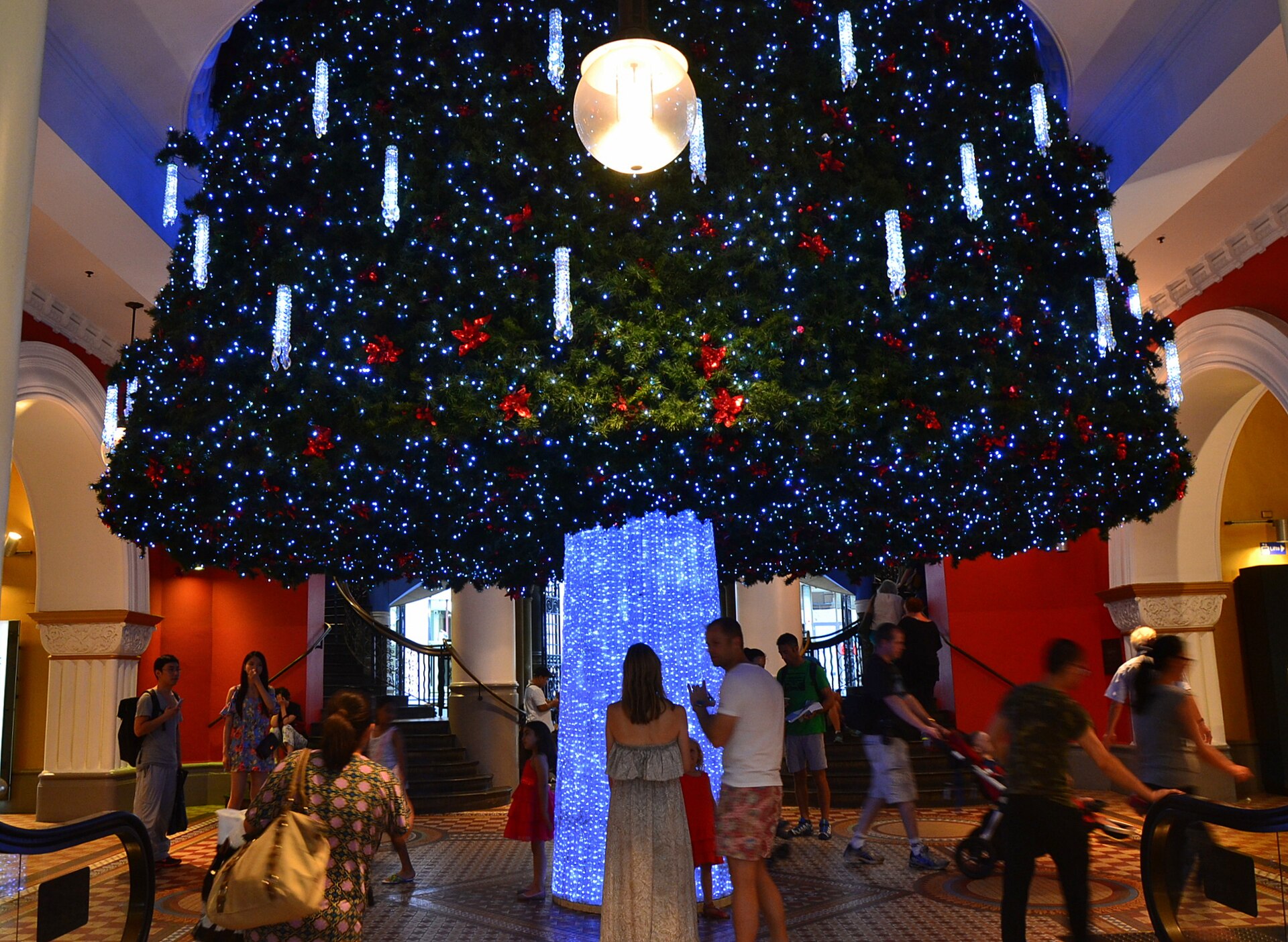 A giant Christmas tree covered in blue and white lights and red ornaments hanging through the atrium of the Queen Victoria Building, with shoppers on the mosaic floor below
