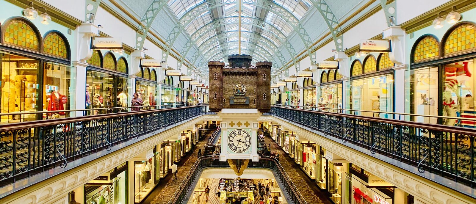 Inside the Queen Victoria Building, looking along the galleried arcade under its arched glass roof to the Royal Clock hanging above the upper floor, with shops on every level and mosaic floors below