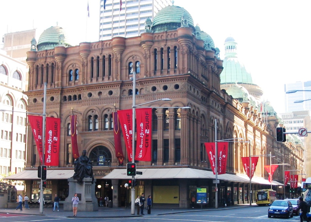The southern end of the Queen Victoria Building on George Street, with its sandstone arches, copper domes and a bronze statue of Queen Victoria on a plinth in front, and red banners on the street poles