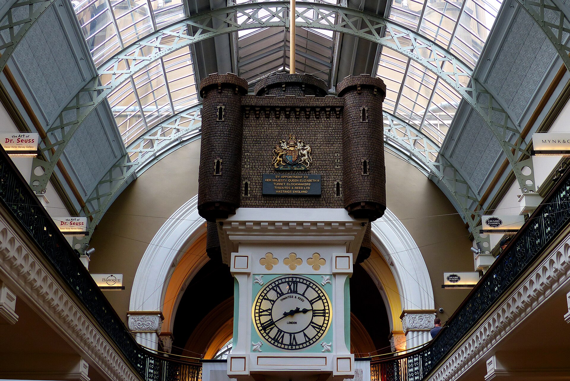 The Royal Clock hanging in the QVB, a model castle with brown turrets, a royal coat of arms and a large clock face in a pale green case, under the arched glass roof