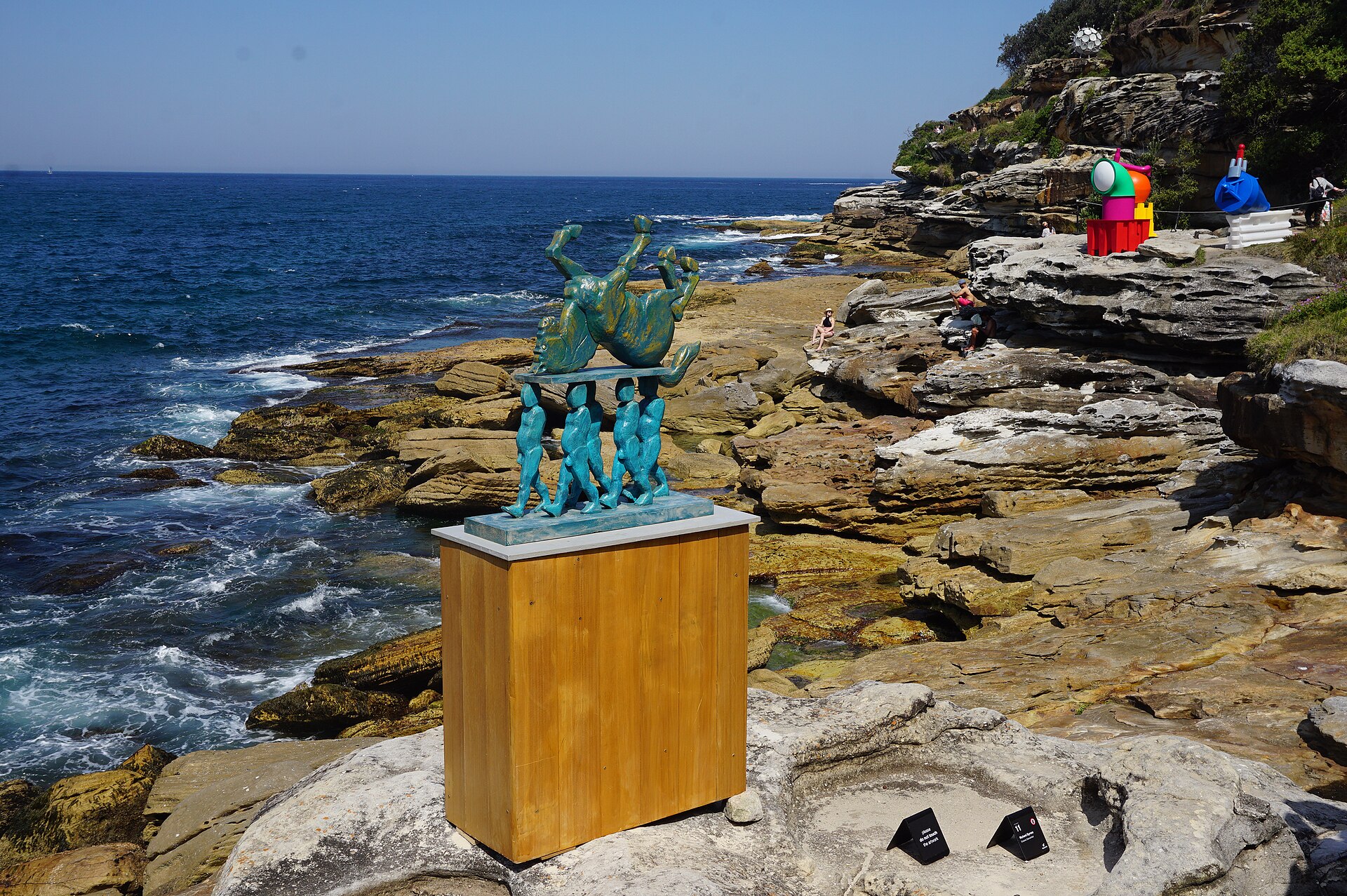 A teal sculpture of a horse carried upside down by a line of figures, on a wooden plinth on the sandstone rocks above the ocean, with more colourful sculptures on the cliffs behind