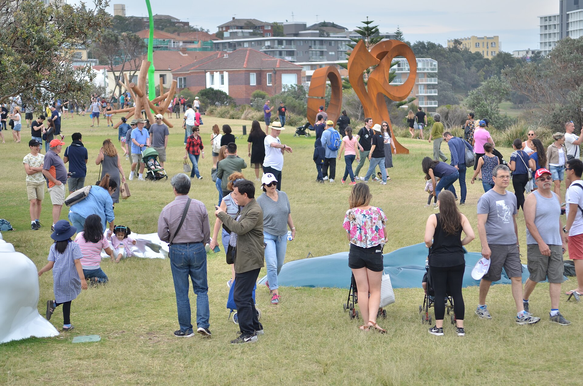 Crowds of visitors walking and picnicking among large rusted steel and timber sculptures on the grass of Marks Park, with apartment blocks behind