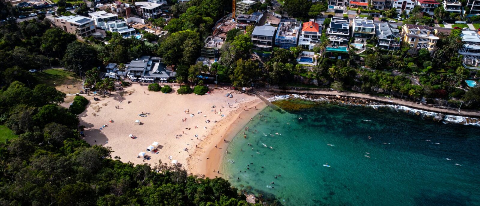 Aerial view of Shelly Beach in Manly, a sheltered sandy cove with clear turquoise water, snorkellers and swimmers, bushland and houses above