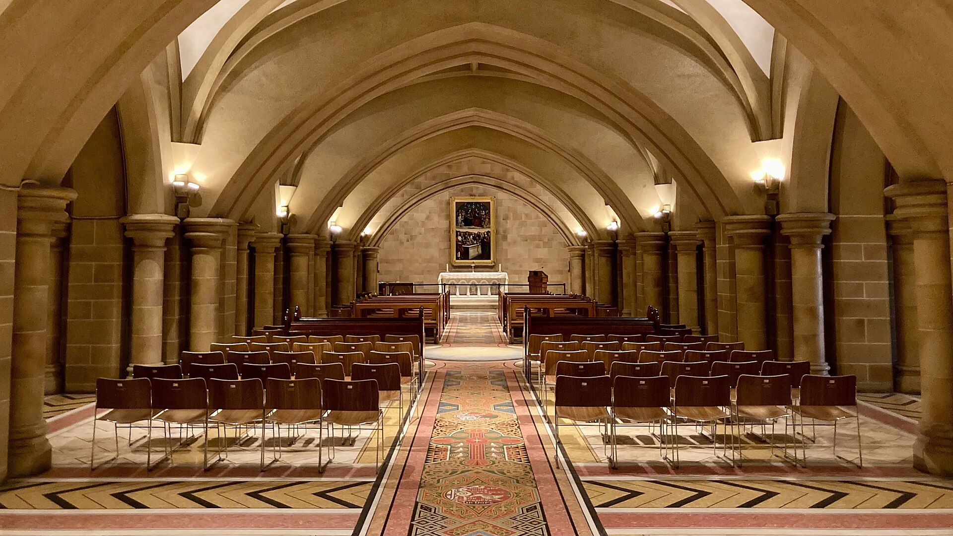 The vaulted sandstone crypt beneath St Mary's Cathedral, with rounded columns, rows of chairs facing a small altar and a painting, and a patterned terrazzo mosaic floor running down the central aisle