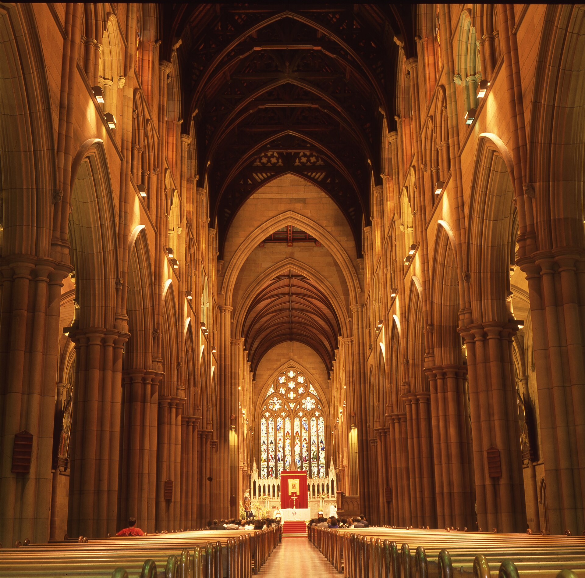 The long nave of St Mary's Cathedral, with sandstone columns and pointed arches lit warm gold, rows of timber pews on both sides of the aisle and the altar under a large stained-glass window at the far end