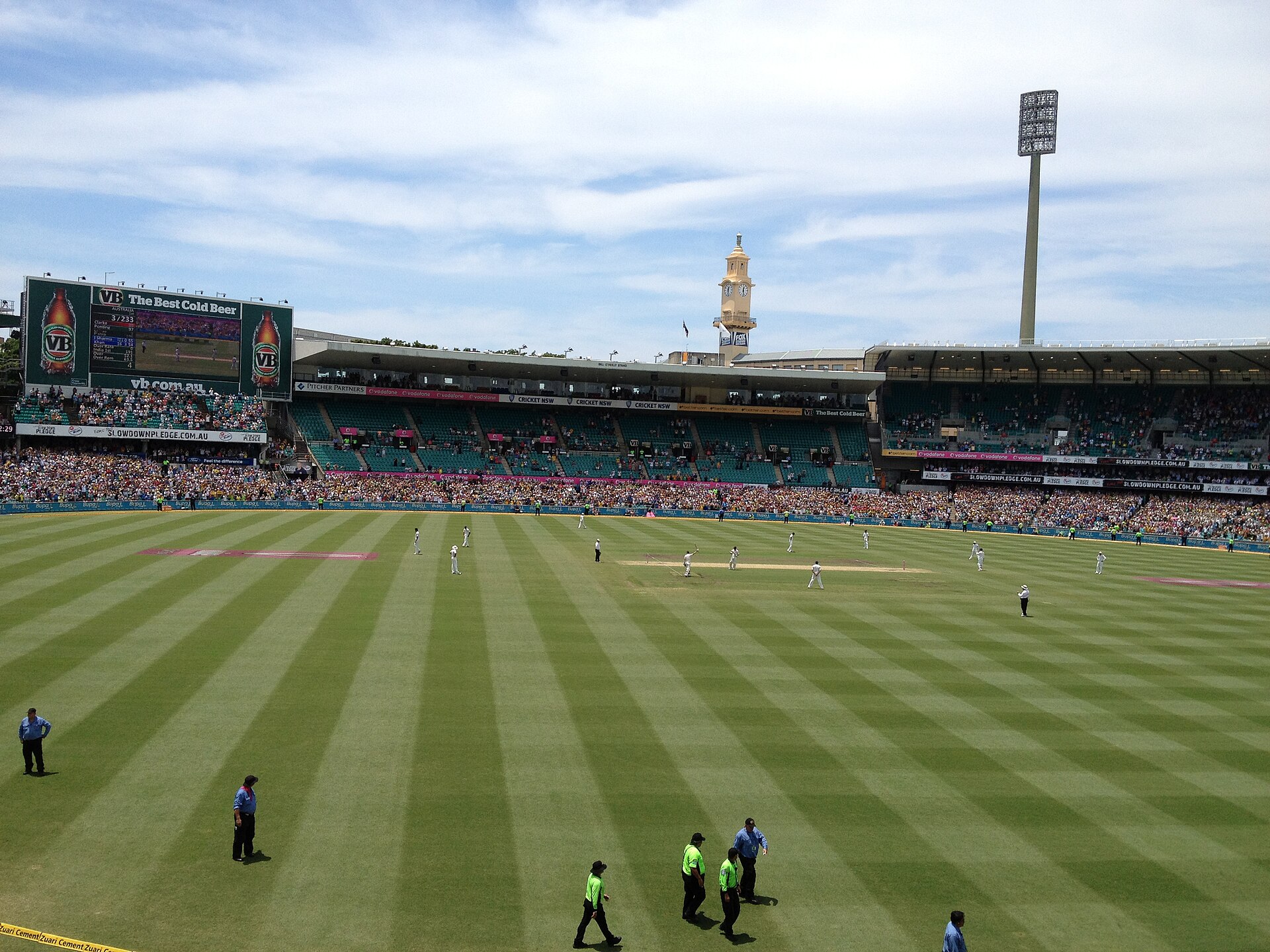 A Test match at the Sydney Cricket Ground on a sunny day, with players on the striped outfield, pink markings on the grass and the historic clock tower above full stands