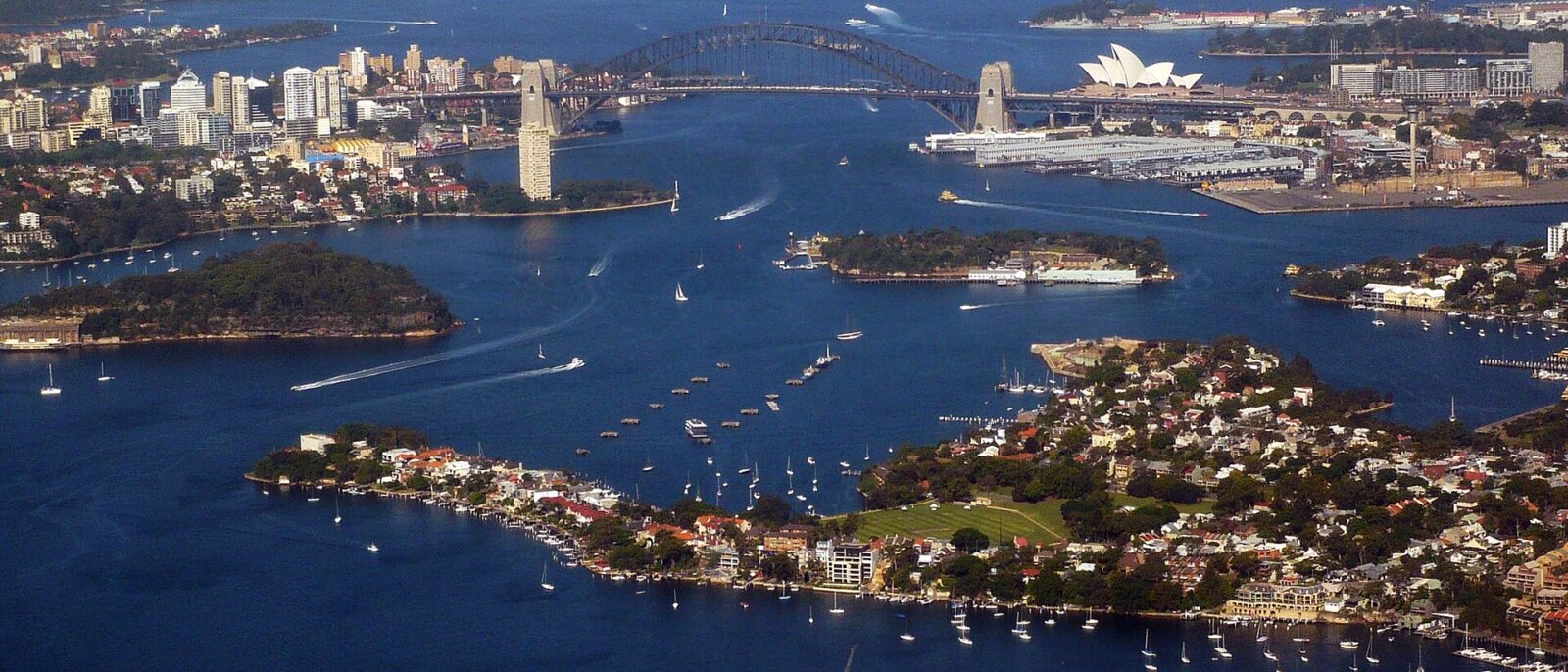 An aerial view of Sydney Harbour with the Harbour Bridge and Opera House in the distance, Goat Island and the Balmain peninsula in the foreground and boats scattered across the blue water