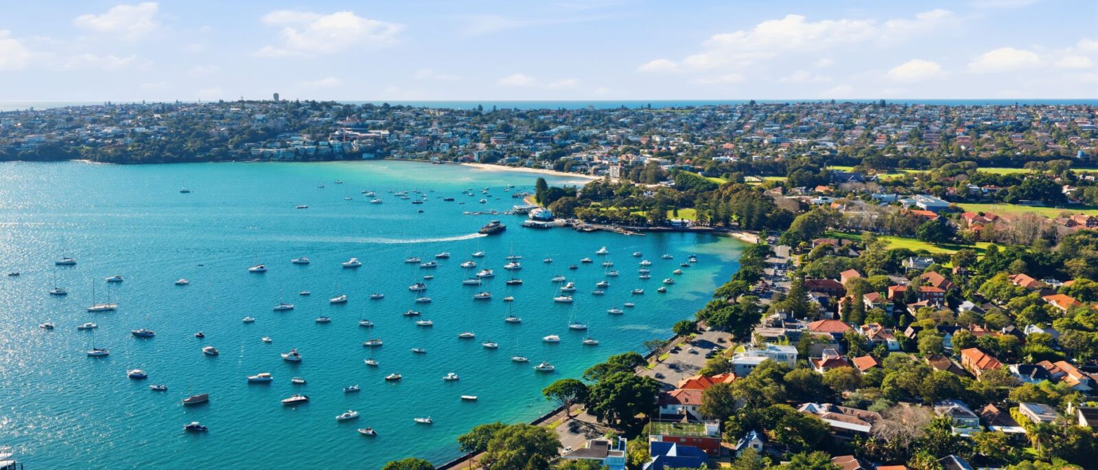 Aerial view of a sheltered bay on Sydney Harbour in the eastern suburbs on a clear day, with moored boats, a ferry leaving the wharf and houses running up to the ocean ridge