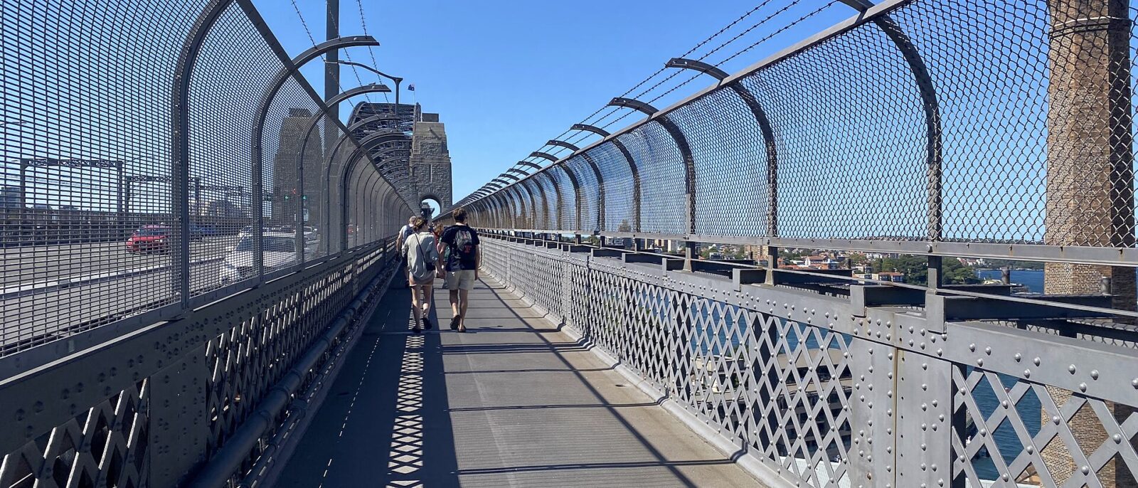 Walkers on the fenced pedestrian footpath of the Sydney Harbour Bridge on a sunny day, with riveted steel railings, the roadway on the left and a granite pylon ahead