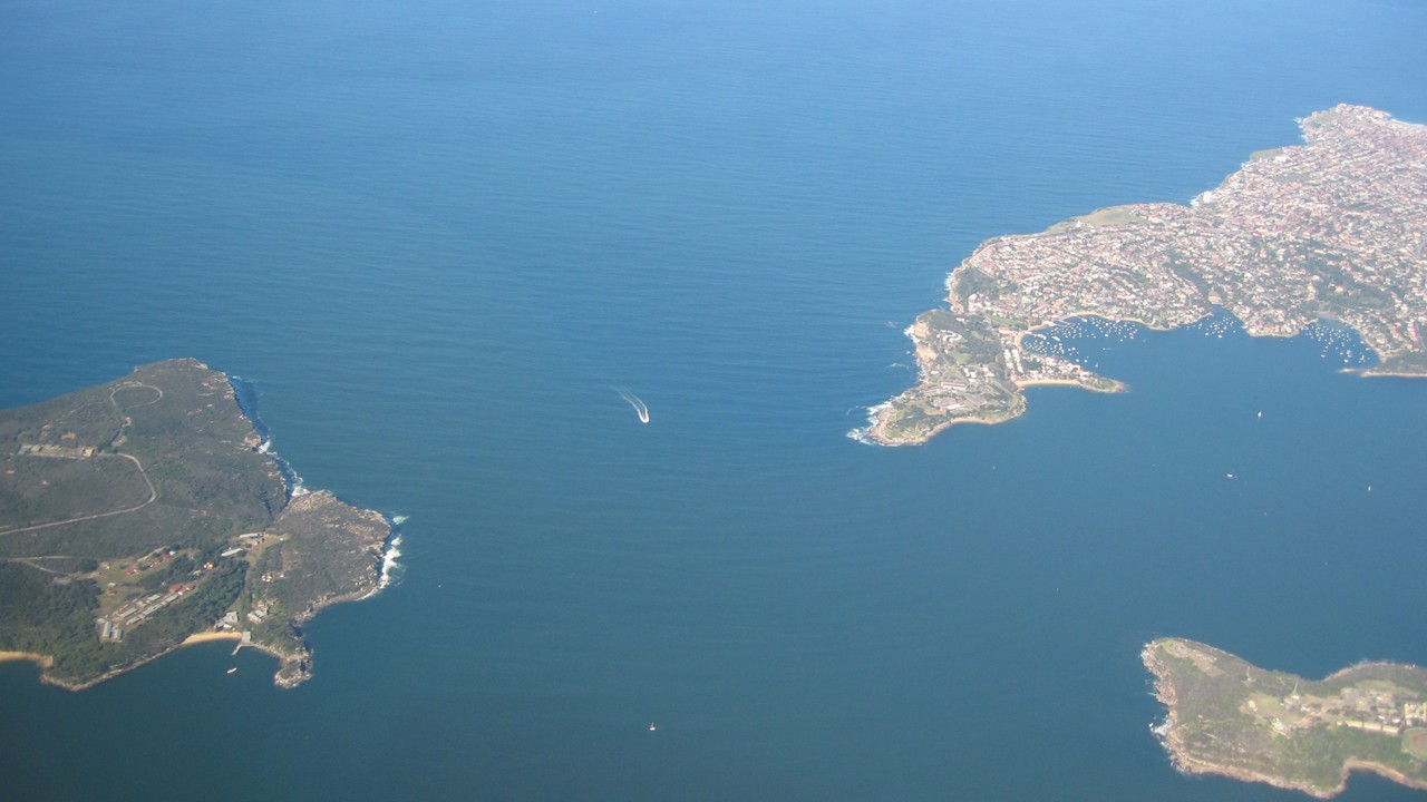 An aerial view of the entrance to Sydney Harbour, with the green plateau of North Head on the left, the suburbs of South Head and Watsons Bay on the right and a boat crossing the deep blue water between them