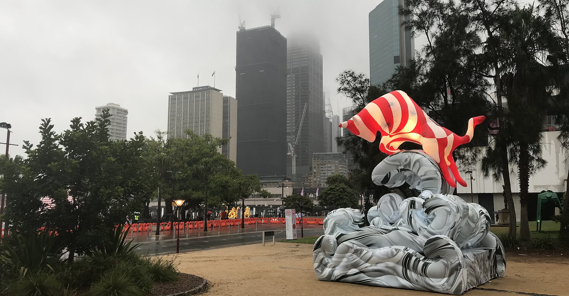 An illuminated red-and-white striped ox lantern rising from a sculpted grey wave at Circular Quay during the Sydney Lunar Festival, with city towers disappearing into low cloud