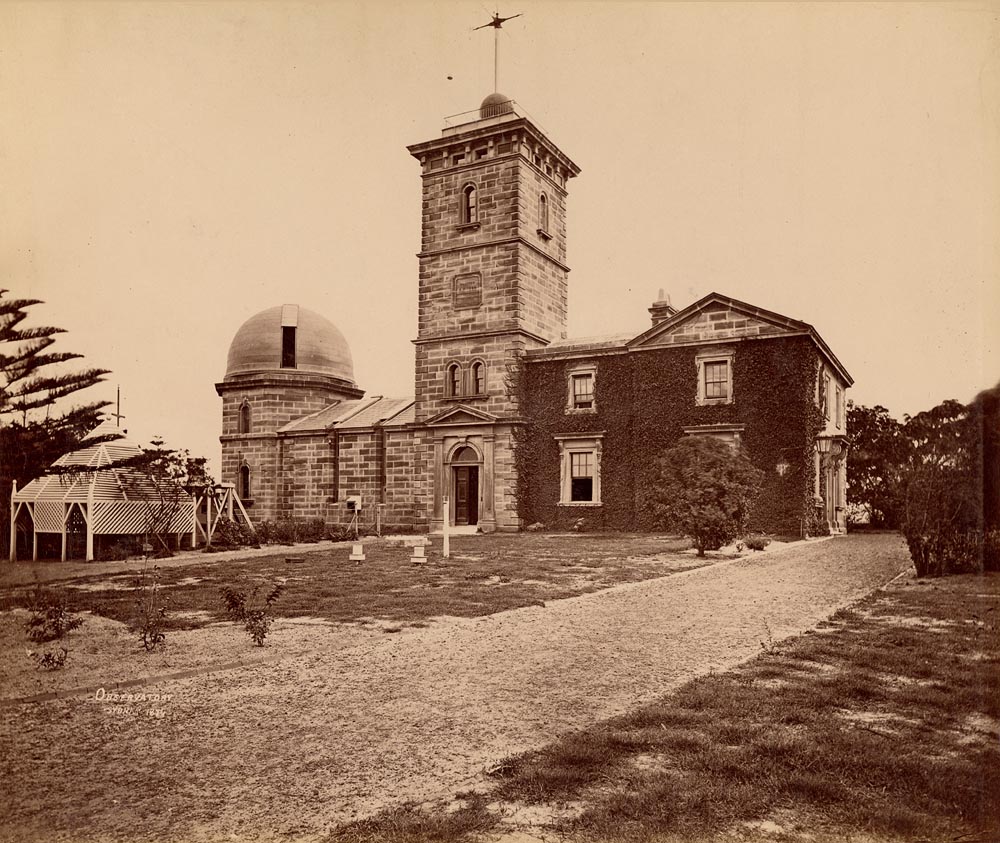 A sepia photograph from 1874 of Sydney Observatory, showing the square sandstone tower with a mast, a small dome and the ivy-covered residence behind a gravel path and young trees