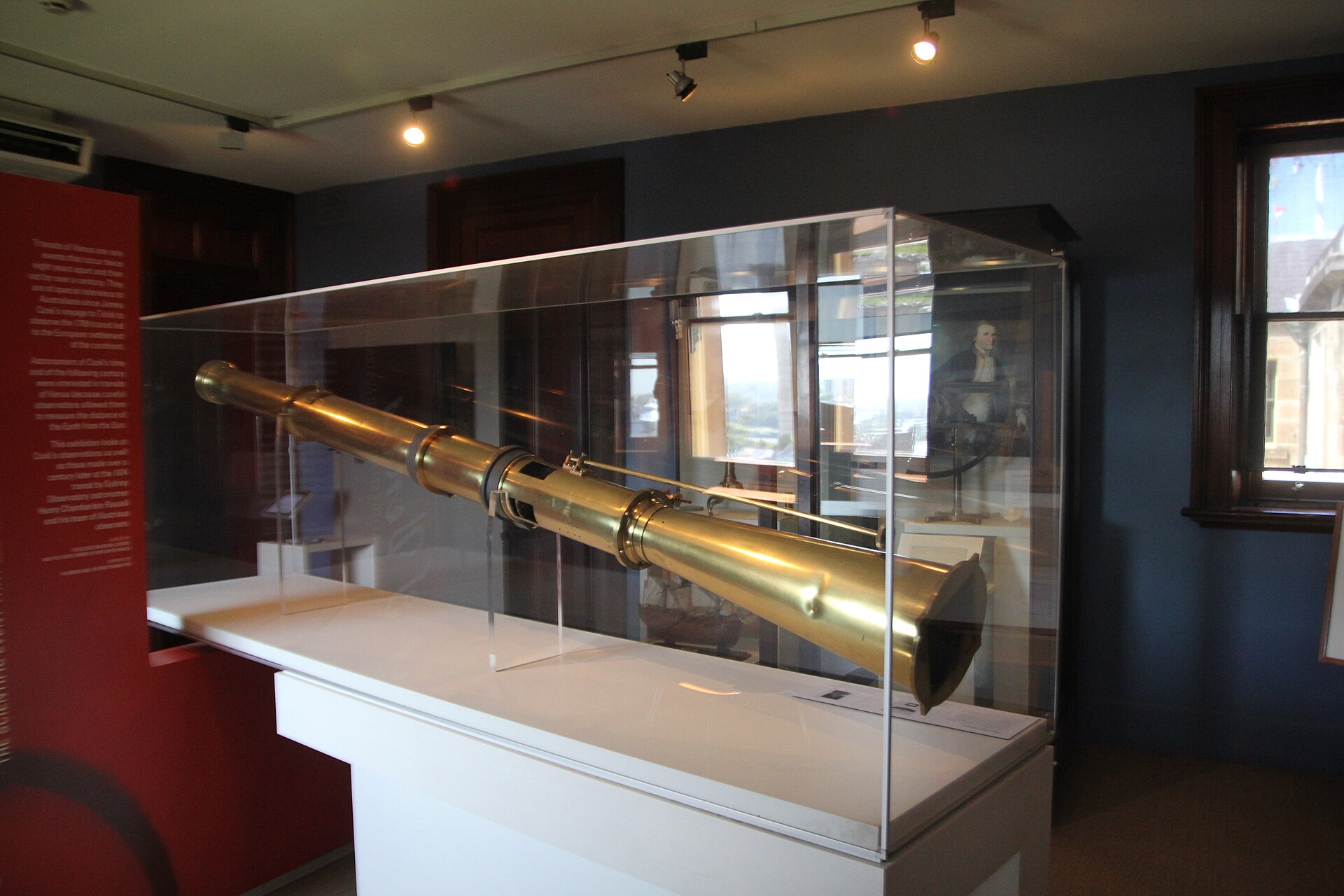 A long brass photoheliograph telescope displayed in a glass case in a museum room with a red wall and a window