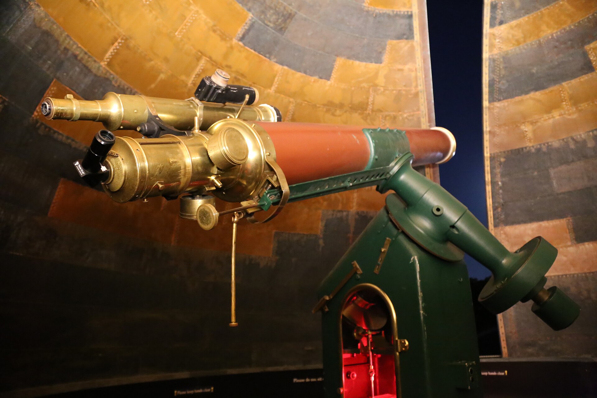 A 19th-century refracting telescope with a long red-brown tube, brass eyepiece fittings and a green iron mounting, inside an observatory dome with an open slit showing the night sky