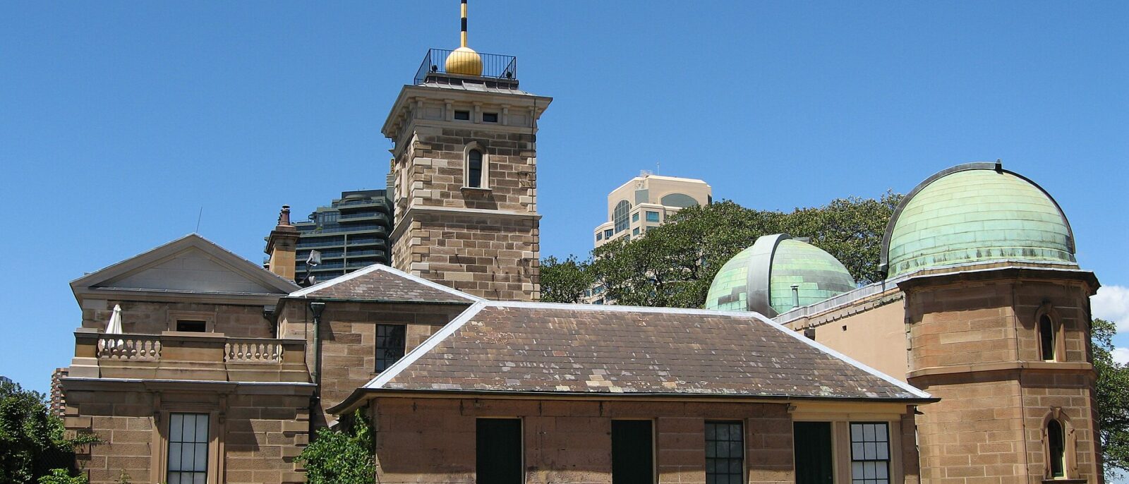 Sydney Observatory under a blue sky: a sandstone building with a square tower topped by a gold time ball and weathervane, and two green copper telescope domes