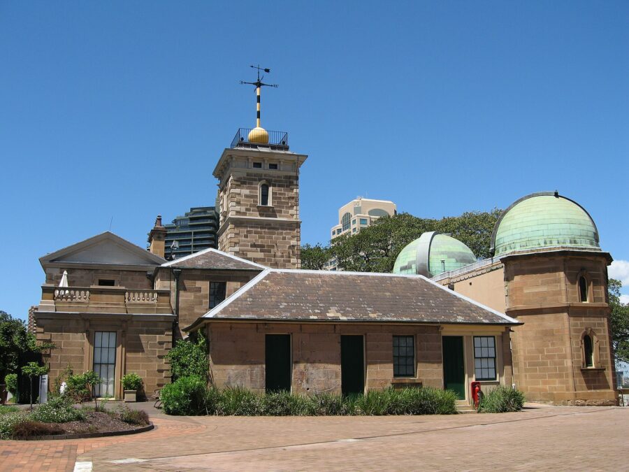 Sydney Observatory under a blue sky: a sandstone building with a square tower topped by a gold time ball and weathervane, and two green copper telescope domes