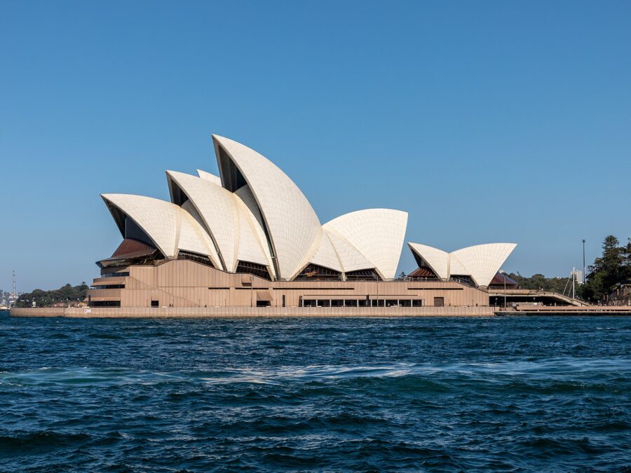 The white sails of the Sydney Opera House seen side-on from the harbour under a clear blue sky, with choppy blue water in the foreground
